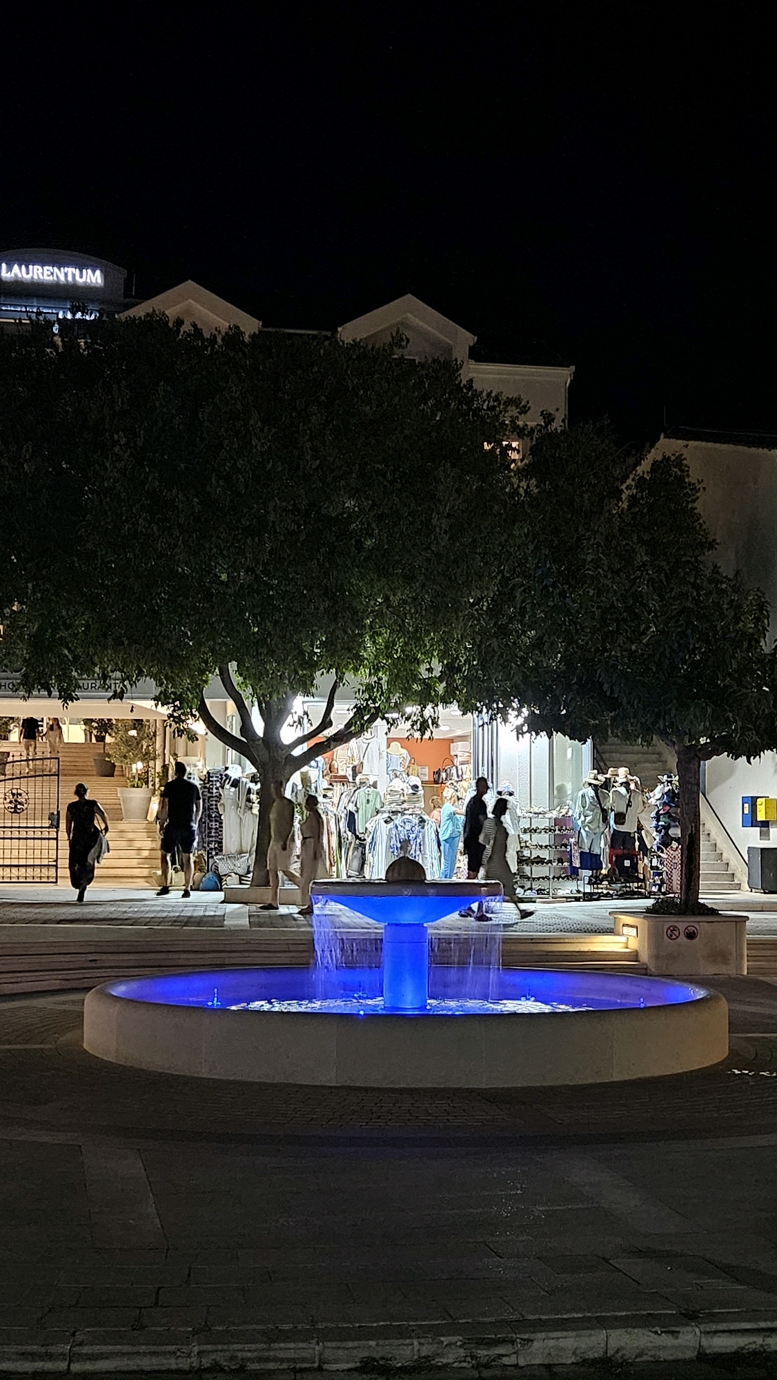 The image depicts a nighttime scene in Općina Tučepi, Croatia, featuring a fountain with blue illumination in the foreground. Behind the fountain, a tree with dark green foliage is prominent. To the right of the tree, a building with shops is visible, displaying various clothing items on racks. Several people are browsing the shops or walking past. In the background, a building with a sign reading "LAURENTUM" is illuminated. The ground is paved with bricks, and the overall atmosphere is that of a commercial area at night.