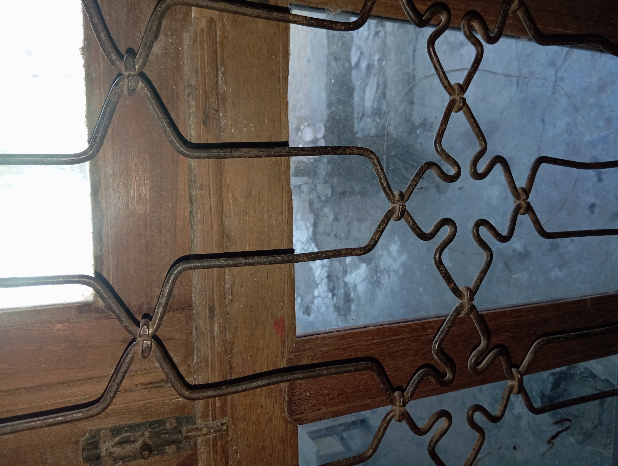 The image shows a close-up view of a window grate in Rejosari, Indonesia. The grate is made of dark, possibly rusted, metal and is attached to a wooden window frame. The metal bars are bent into a decorative pattern of interconnected diamond shapes and zigzags. The window itself appears to be glass, and through it, a blurry, textured surface that could be a wall or floor is visible. The overall impression is of an aged and somewhat weathered architectural feature.