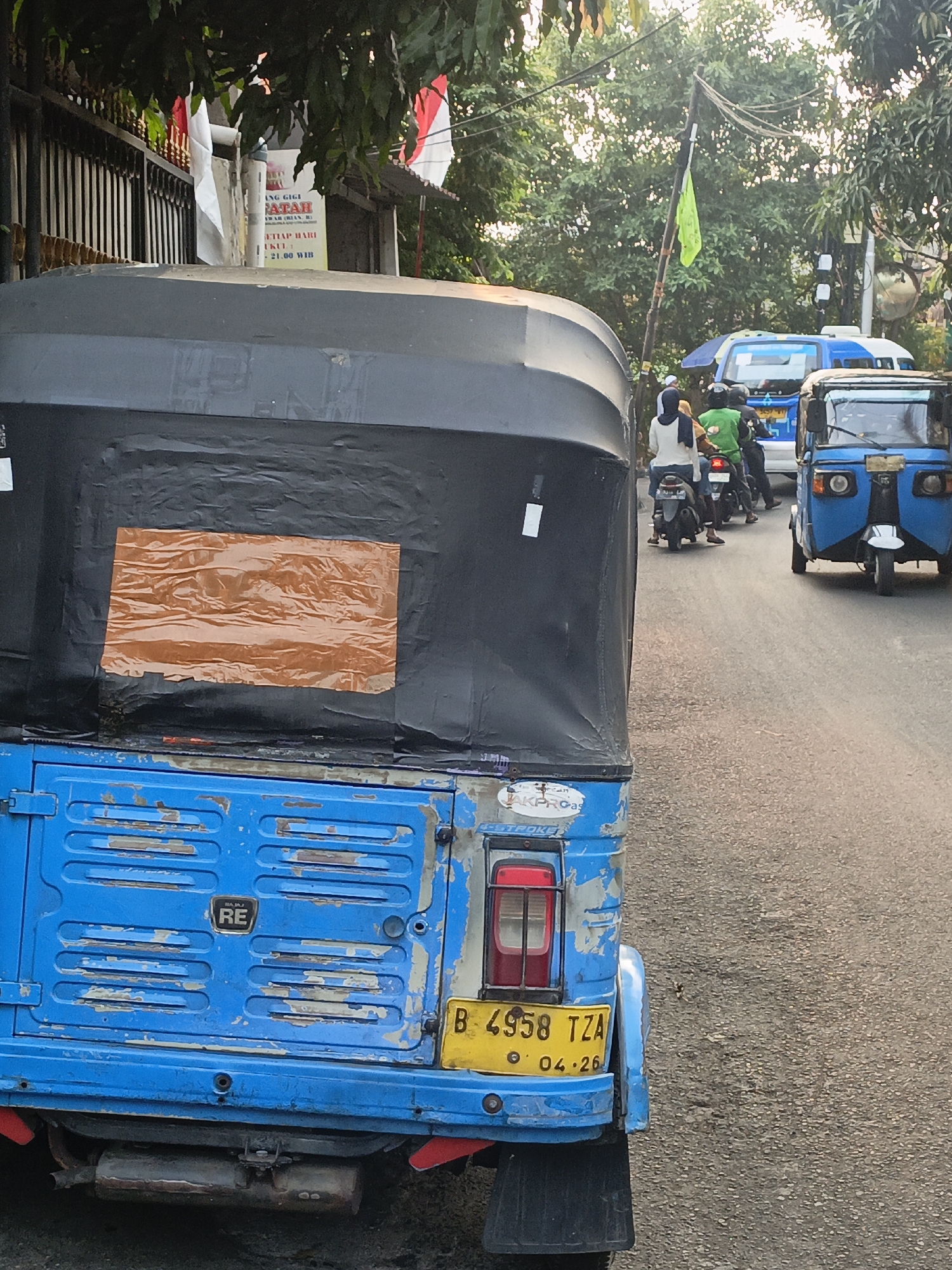 The image captures a street scene in East Jakarta, Indonesia, with a focus on a blue auto-rickshaw in the foreground. The auto-rickshaw is partially obscured by a larger vehicle, but its weathered blue paint, faded from use, is visible. A large orange rectangle, possibly a repair patch or decoration, is affixed to the black canopy of the auto-rickshaw. The vehicle's rear features a license plate that reads "B 4958 TZA" and a date stamp "04.26".
Behind the auto-rickshaw, other vehicles are present, including a blue and white van and another auto-rickshaw. Several people are visible on motorbikes, some wearing helmets. A lime green flag flutters from a pole in the background, amidst lush green trees and overhead electrical wires. The setting appears to be a moderately busy street, possibly during daylight hours, with evidence of a commercial establishment on the left side, indicated by signage and Indonesian flags. The overall impression is that of a typical urban streetscape in Indonesia.