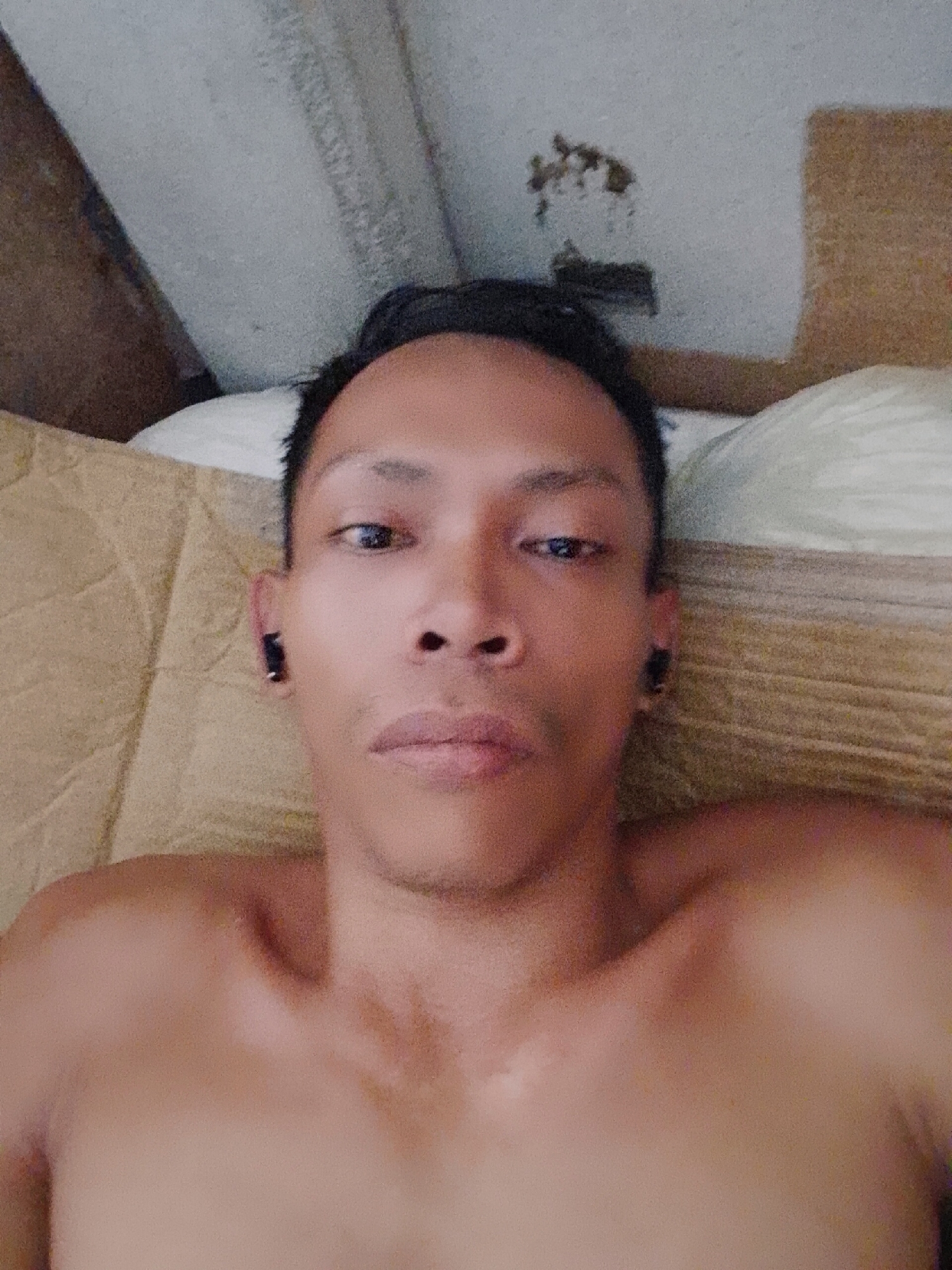 A shirtless person with short black hair, dark eyes, and visible collarbones is lying down, presumably on a bed, in an indoor environment. They appear to be wearing black earbuds. The background shows a wall or ceiling, possibly with some damaged areas or stains. The location is noted as Bukit Ketok, Indonesia, but there is no visual information to verify this.