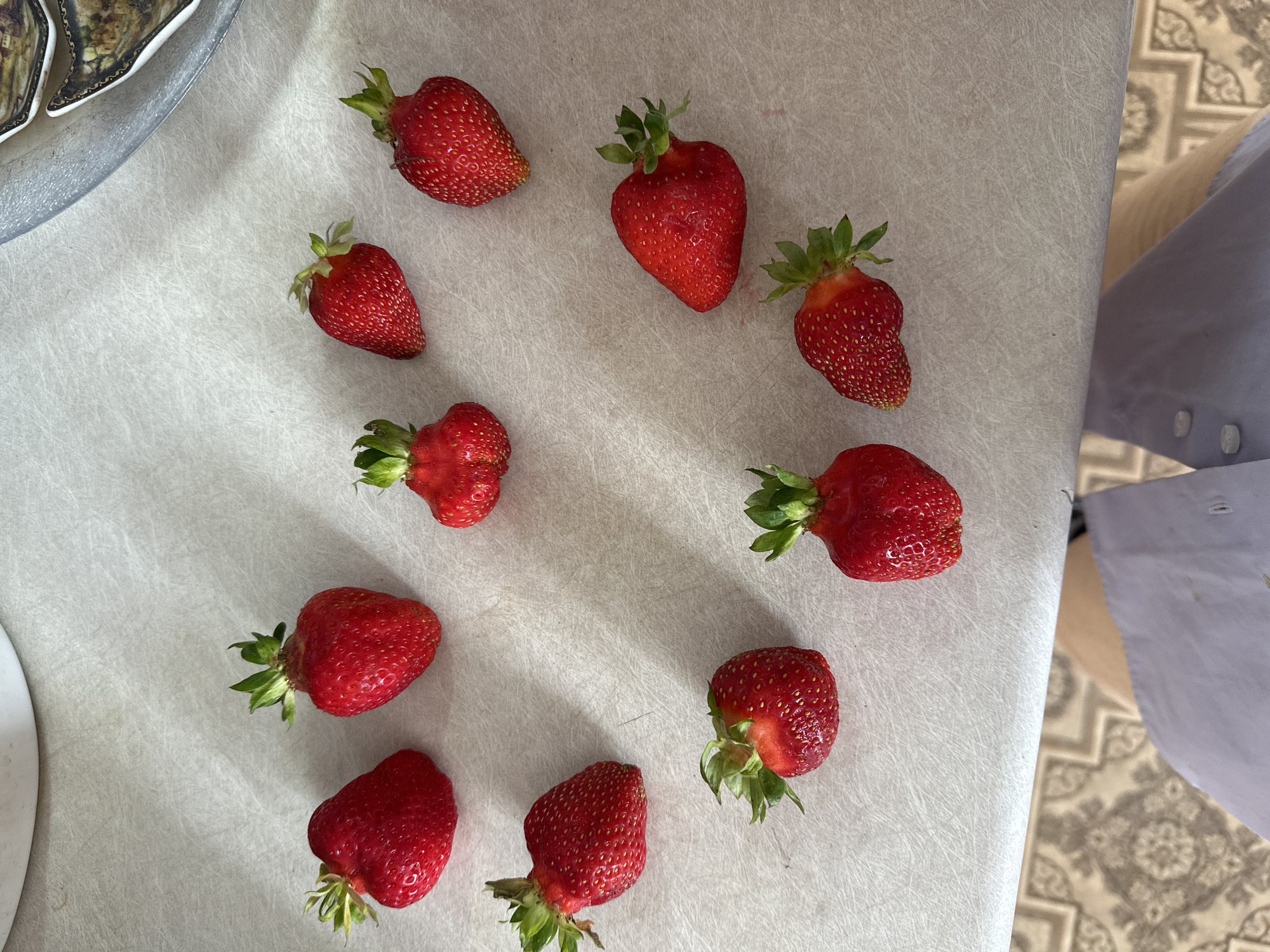 Ten strawberries are arranged in a rough heart shape on a light-colored, textured countertop. The background includes a white bowl or container at the top, a silver platter with decorative plates on the right, and a patterned carpet on the lower portion. The lighting appears to be natural, coming from the right, casting slight shadows. The image was taken in Yaroslavka, Russia.