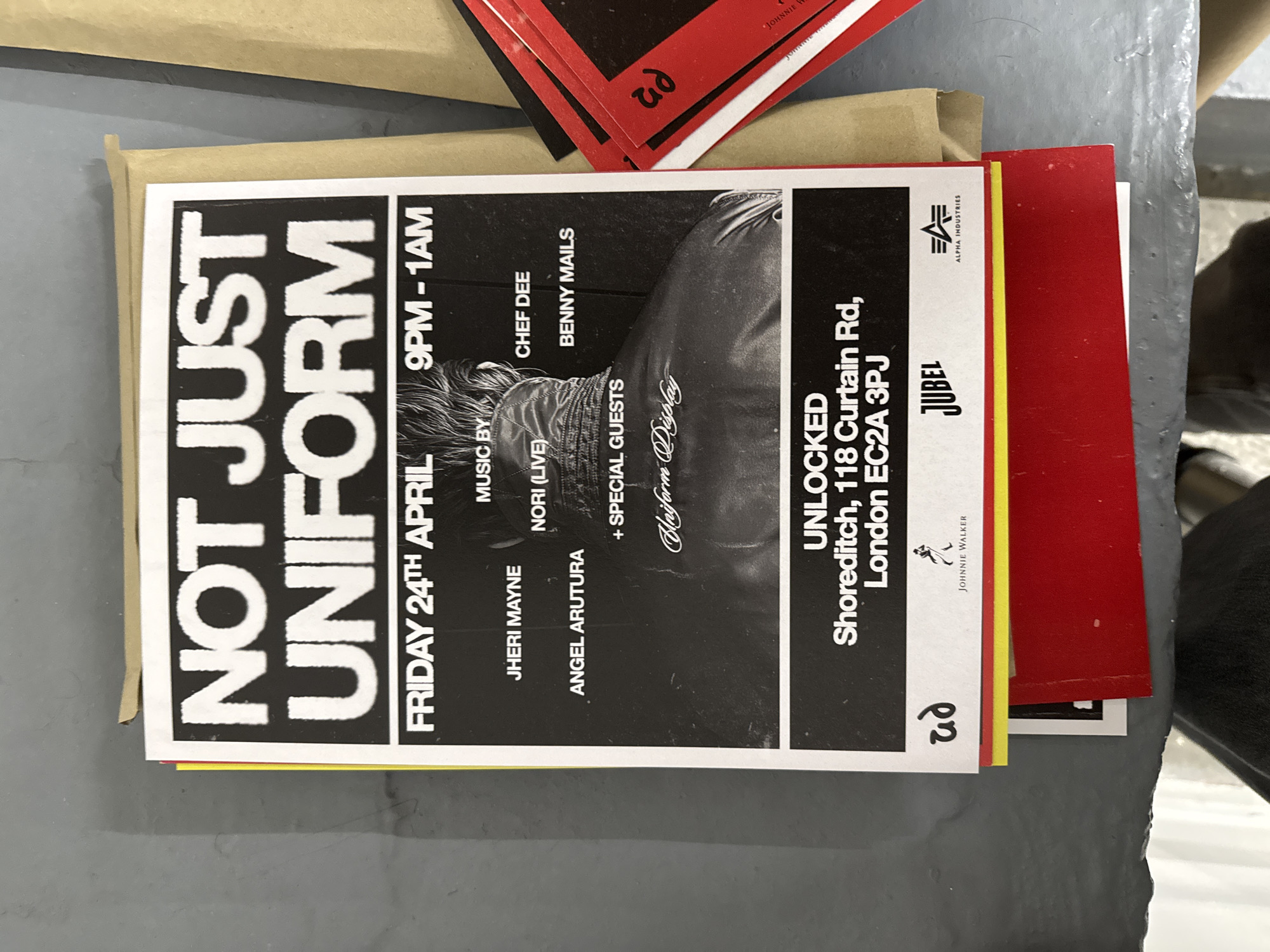 An event flyer is prominently displayed in the center of the image, lying horizontally on a grey, textured surface. To the left, a piece of light brown cardboard or paper is visible. To the right, several red cards or smaller flyers are stacked, with some partially obscured.
The main flyer is for an event titled "NOT JUST UNIFORM" and features a striking black and white design. The date and time are "FRIDAY 24TH APRIL" from "9PM - 1AM". A list of performers or DJs includes "JHERI MAYNE", "ANGEL ARUTURA", "NORI (LIVE)", "CHEF DEE", and "BENNY MAILS", along with "+ SPECIAL GUESTS Blayform Display".
The event's location is specified as "UNLOCKED Shoreditch, 118 Curtain Rd, London EC2A 3PJ". Several logos are visible: "ALPHA INDUSTRIES" in the top right, "JUBEL" and "JOHNNIE WALKER" towards the bottom right, and a stylized "ad" logo appears twice, once on the flyer itself and once on one of the red cards.
The central visual element on the flyer is a black and white photograph of an individual from the shoulders up, turned away from the viewer, showing textured hair and a jacket. A yellow vertical stripe runs along the right edge of the flyer. The overall impression is an advertisement for a music or club event in London, United Kingdom.