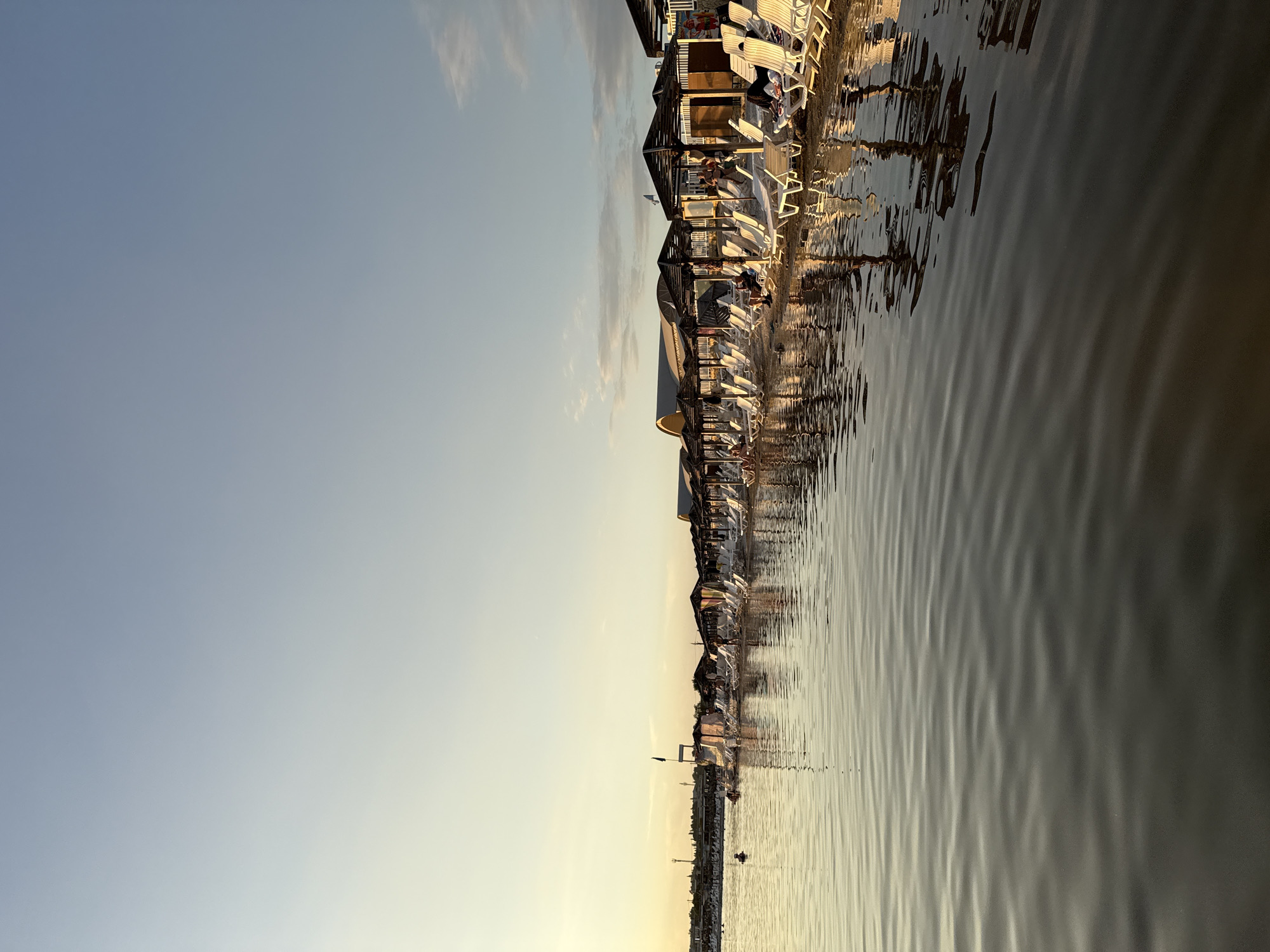 The image depicts a beach scene in Sol-Iletsk, Russia, during what appears to be late afternoon or early evening, indicated by the soft, golden light and long shadows. The foreground is dominated by calm, rippling water, reflecting the sky and the beach scene above. Along the shoreline, a row of white lounge chairs are set up under dark, angled umbrellas. Several people are visible, some reclining on the chairs, others walking or standing. A lifeguard stand with a black flag is positioned further down the beach. In the distance, structures and trees can be seen, suggesting a developed area beyond the beach. The sky is a pale blue, with some wispy clouds near the horizon, transitioning to a lighter, yellowish hue as it meets the distant land. The overall impression is one of a relaxed, recreational environment.