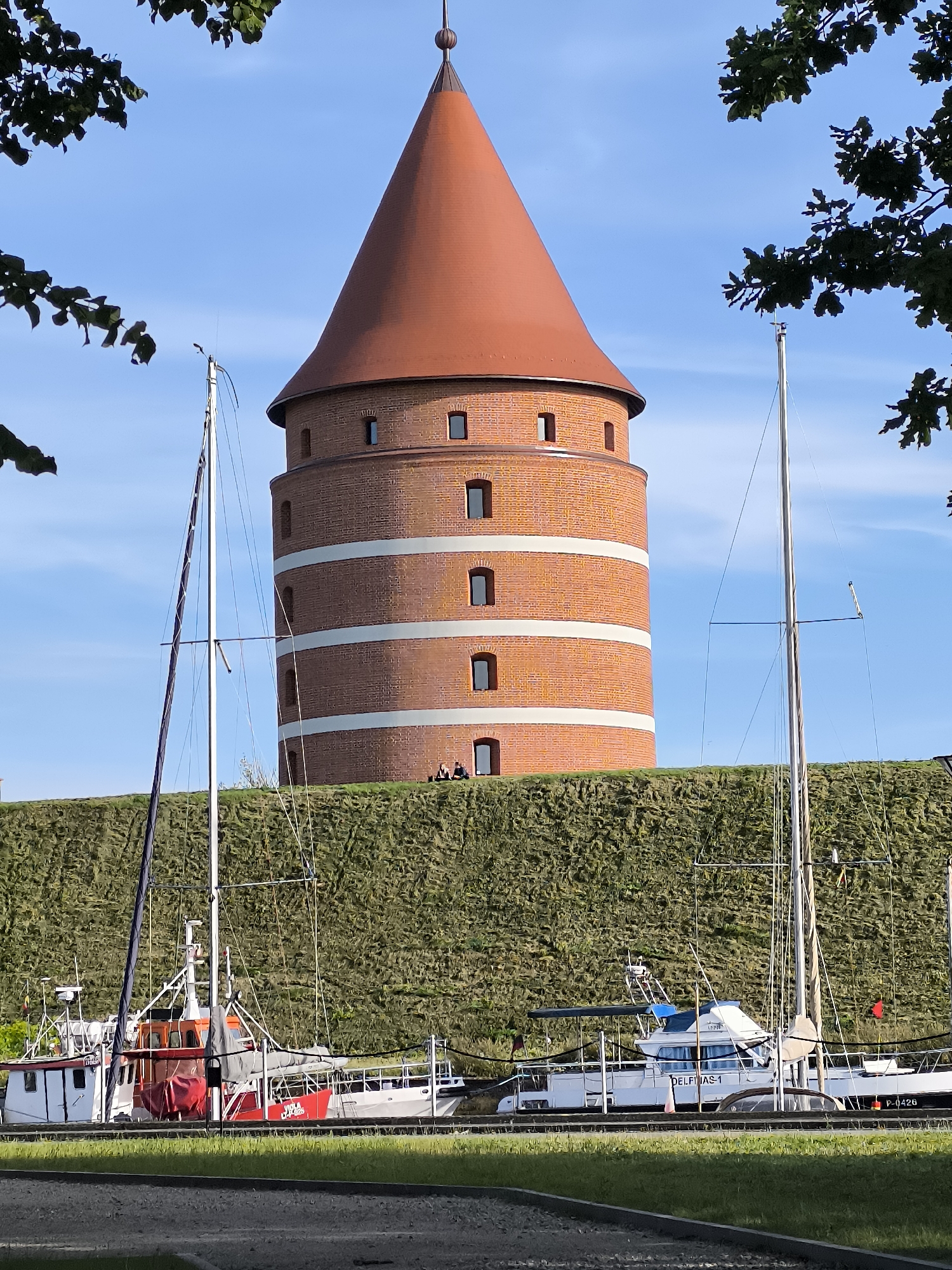 The image captures a scene in Klaipėda, Lithuania, featuring the iconic red brick tower of the Klaipėda Old Post Office, also known as the "Old Mail Tower." The tower, with its conical, red-tiled roof, is the central subject, standing tall against a clear blue sky. It is surrounded by a lush green embankment. In the foreground, several boats are moored, including a white and red fishing vessel with the inscription "VIOLA JUNA 1925" on its side, and a larger white yacht named "DELFINAS-1". The scene is framed by the dark leaves of trees on the left and right, suggesting it is a sunny day with good visibility. Two individuals are visible sitting on the embankment near the base of the tower. The overall impression is one of a historical landmark situated in a maritime environment.