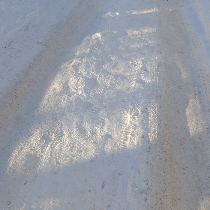 The image shows a snow-covered surface marked with various footprints and tire tracks. The snow appears to be compacted in the areas where vehicles have passed, leaving distinct impressions. Footprints are scattered across the surface, adding to the textured appearance. The lighting suggests a bright, sunny day, with shadows subtly enhancing the contours of the snow and the tracks. The overall scene presents a wintery, lived-in landscape.