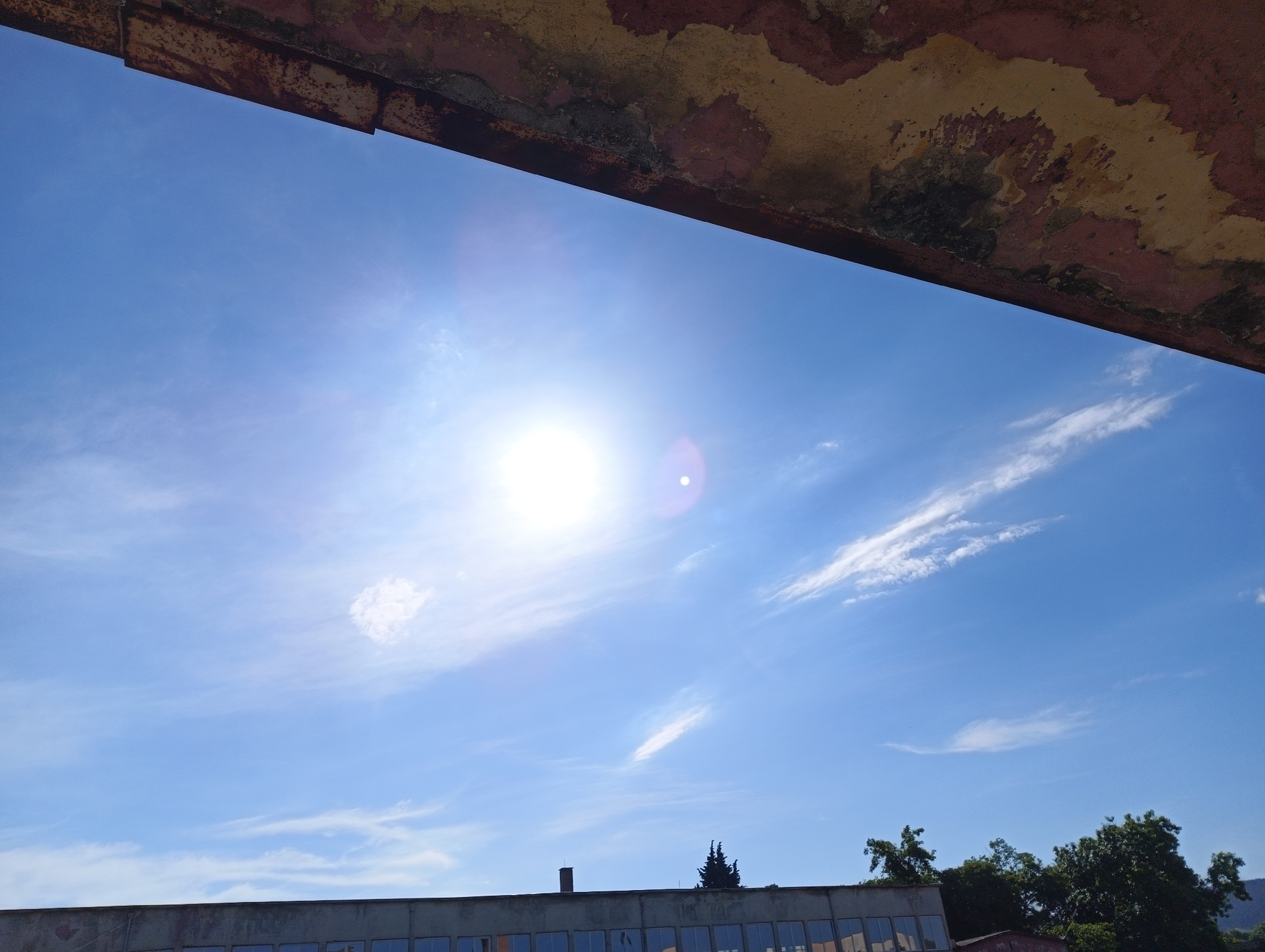 A low-angle shot captures a clear blue sky with wispy clouds, partially framed by what appears to be the underside of a deteriorating building or structure at the top of the frame. The sky is dominated by a bright sun, causing lens flare in the center. At the bottom of the frame, the upper portion of a building is visible, including a line of windows, and some trees are seen to the right. The location appears to be Unknown, Unknown.