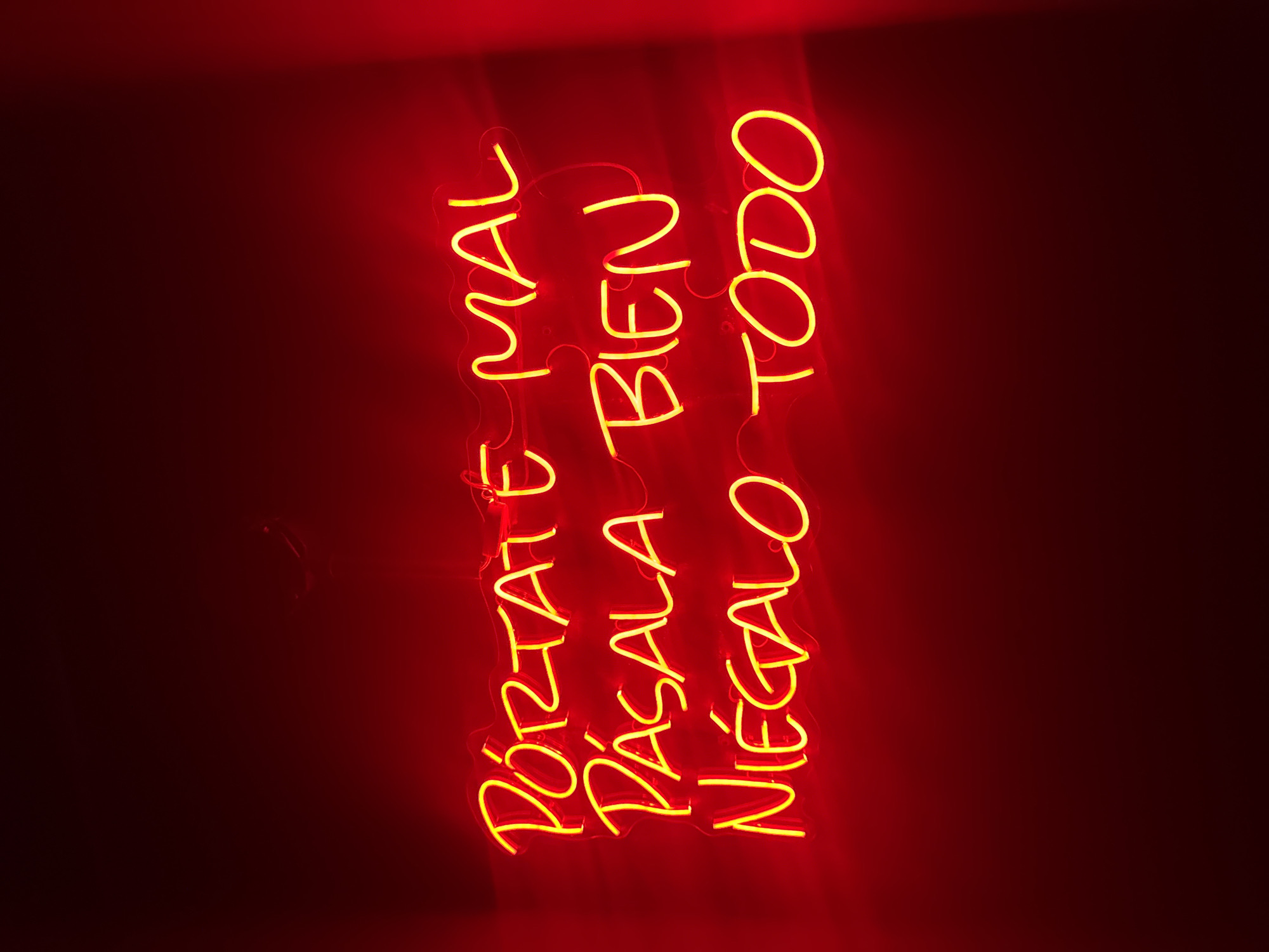 A neon sign, illuminated in a vivid yellow-orange, is set against a deep red background. The words are written in a looping, cursive style, stacked vertically. The sign appears to display three distinct phrases: "Pórtate Mal", "Pásala Bien", and "Niégalo Todo". The setting or location is not evident in this image, therefore, providing the location is not possible.