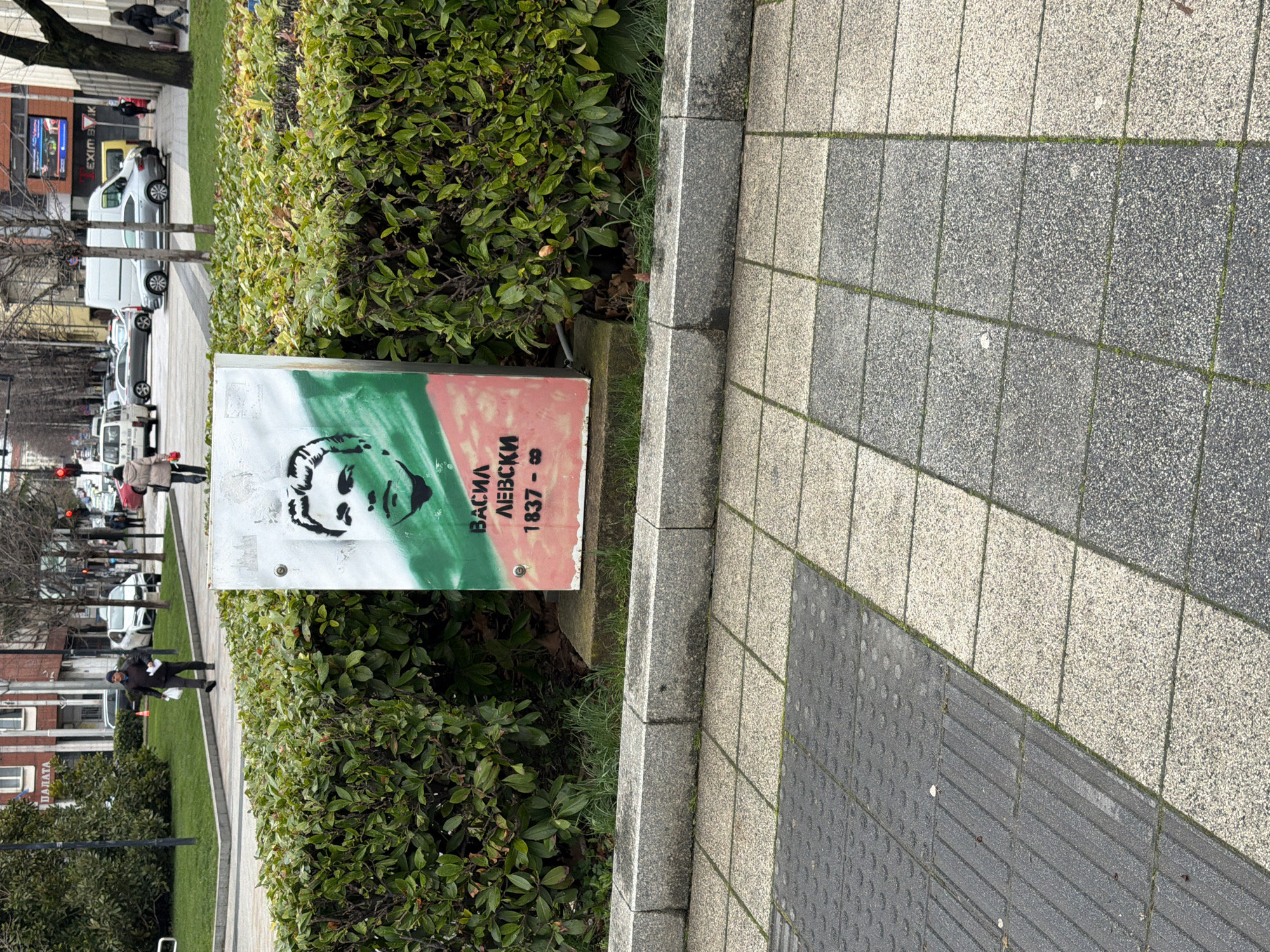 The image captures a street scene in Pleven, Bulgaria. A white metal box, decorated with a stencil portrait of Vasil Levski and his birth year of 1837 in Bulgarian, sits between a hedge of greenery and a grey-and-white paved sidewalk. The portrait is colored with green and pink stripes of the Bulgarian flag. In the background, blurred figures of pedestrians walk along the sidewalk, and cars are parked along the street. Trees without leaves line the street, and buildings are visible in the distance.