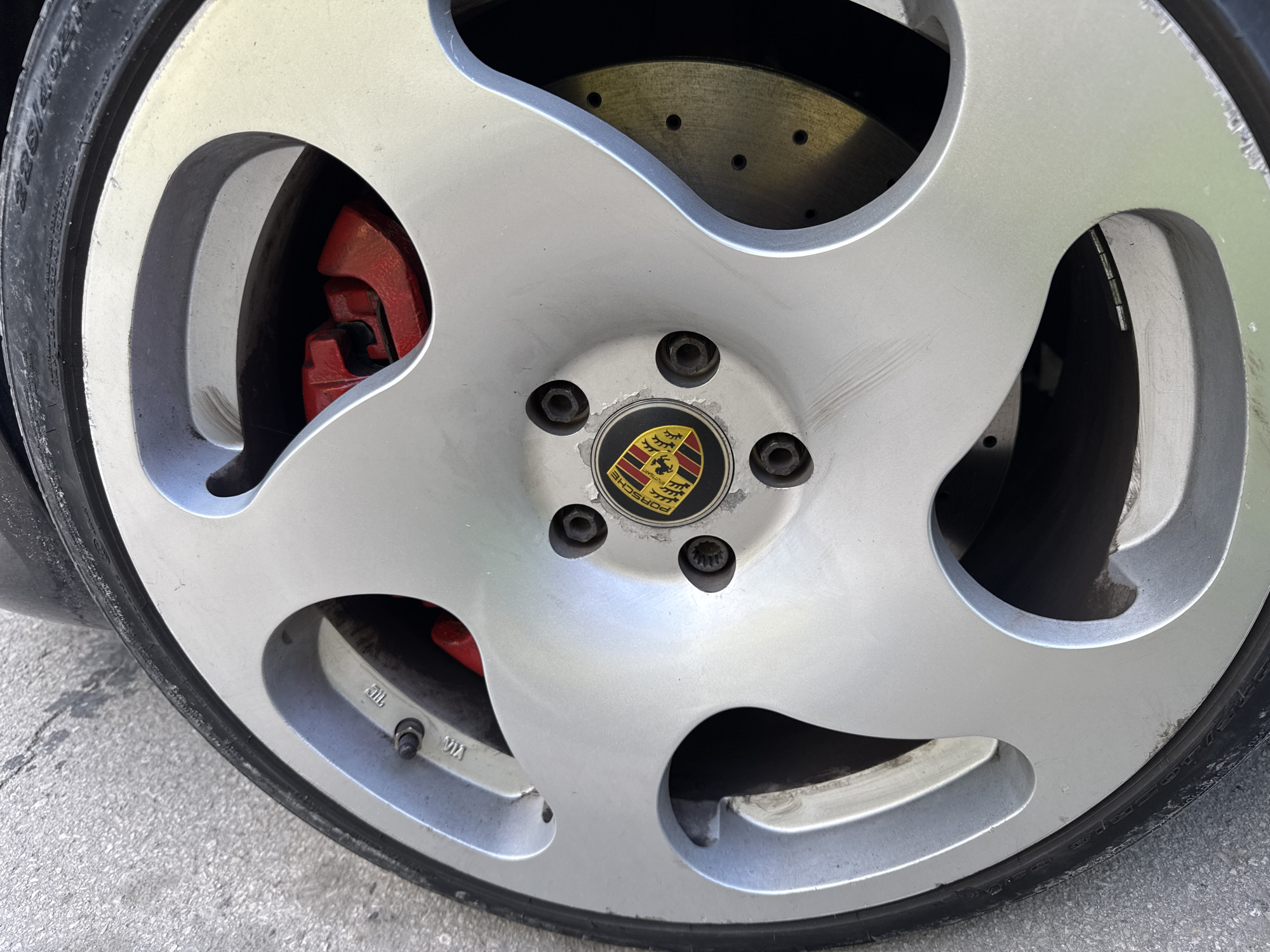 A close-up, eye-level shot focuses on the front passenger-side wheel of a silver Porsche with red brake calipers. The wheel is a silver alloy with a multi-spoke design, featuring the Porsche crest in the center. The tire is black and has tire pressure information visible. The background consists of a grey concrete surface. The image was taken outdoors during daylight. The specific location in Varna, Bulgaria, is not ascertainable from the visual information provided.