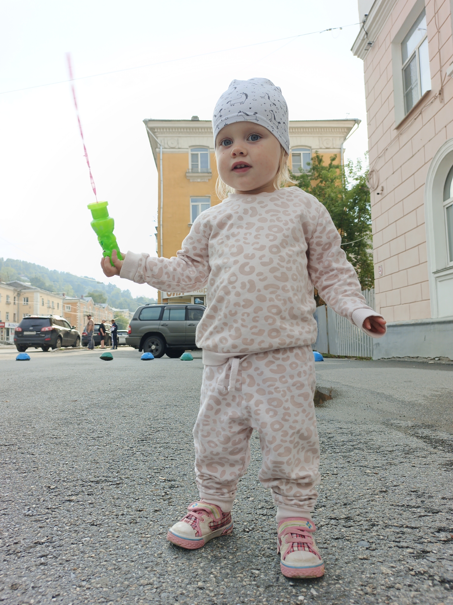 A young Caucasian girl, estimated to be between 2-3 years old, stands in a paved area outdoors, holding a green bubble wand with a pink string. She is wearing a light-colored beanie with a pattern of small dark animal figures, a long-sleeved sweatshirt and matching pants, both with a light pink leopard print. Her feet are clad in pink and white sneakers with laces. The background shows a street with parked cars, including a black SUV and a dark green SUV. In the distance, buildings with yellow and pink facades are visible, along with trees and a hilly landscape. The scene appears to be in an urban environment. The presence of a building with signage in Russian, potentially "Zlatoust Metallurgical Plant," suggests the location is Zlatoust, Russia. The ground is wet in places, indicating recent rain.