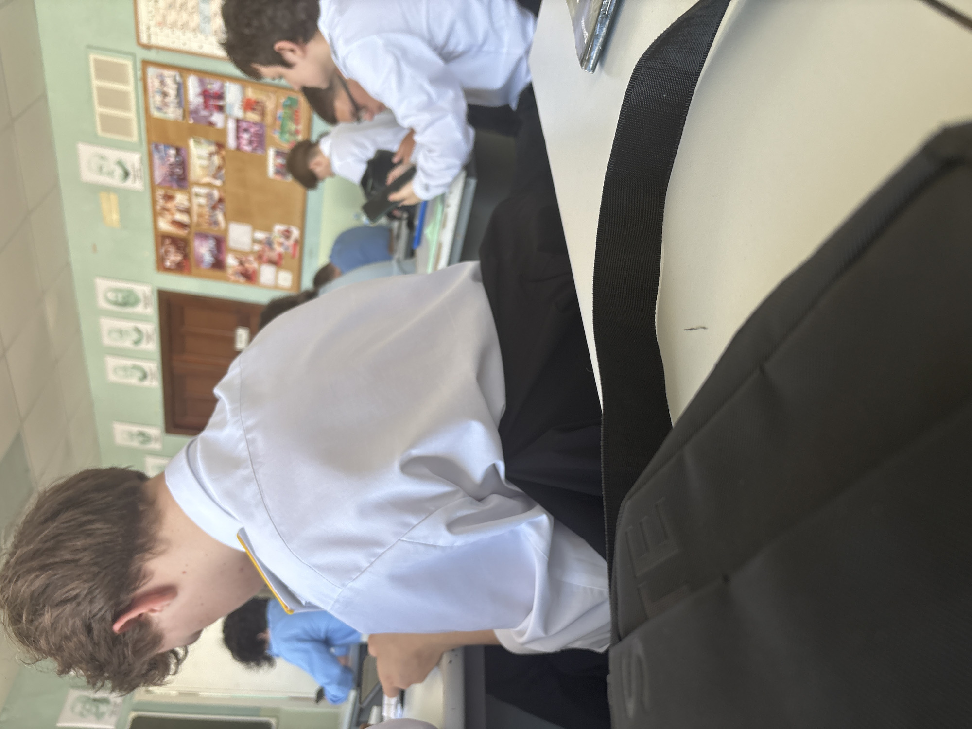 The image depicts an indoor scene, likely a classroom or educational setting, viewed from a slightly elevated perspective. In the immediate foreground, to the left, is the back of a young person's head with short, brown, wavy hair, and their left shoulder, wearing a white collared shirt with a yellow trim detail. To the right, a black bag, possibly a shoulder bag or backpack, rests on a white desk or surface. The bag has a textured surface, and the brand "Ulefone" is partially embossed on its side. A black strap for the bag runs diagonally across the white surface.
In the midground, several other young people, presumably students, are visible. They are also wearing white shirts, suggesting a uniform. One student, seen from behind, is leaning over a desk. Another student, further back and to the right, is wearing a light blue top. The students appear to be engaged in typical classroom activities, either listening, working, or interacting.
The background features a light green wall. On this wall, a large cork noticeboard is prominently displayed, filled with numerous papers, photos, and colorful prints. Many of these appear to be images of characters, possibly from anime or manga, suggesting artistic projects or personal interests displayed by students. Below and to the left of the noticeboard, several framed or pinned white sheets of paper with green sketches of faces or figures are arranged vertically. A wooden door is partially visible in the far background on the left side.
The setting indicates a school environment during the daytime, though the specific time of day is not discernible beyond general daylight. The overall mood appears neutral, characteristic of a routine classroom session.