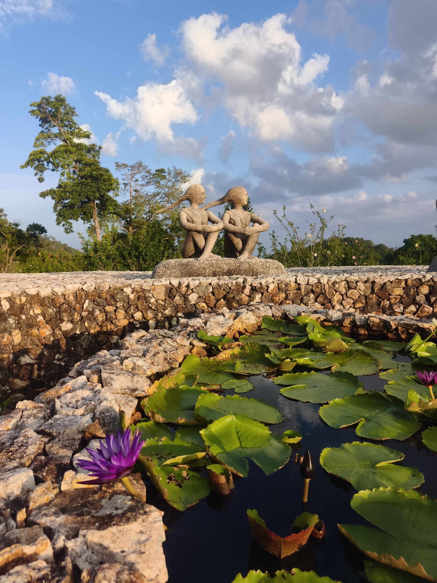 This is a high-angle medium shot of a pond with lily pads and flowers in the foreground, bordered by a stone wall. Behind the wall, two stone statues of young people with long hair are seated on a rock. The background consists of trees and a blue sky with white clouds. The scene is outdoors and appears to be taken during the day. This image is likely located in Ko Samui District, Thailand, given its tropical vegetation and the presence of lotus flowers.
