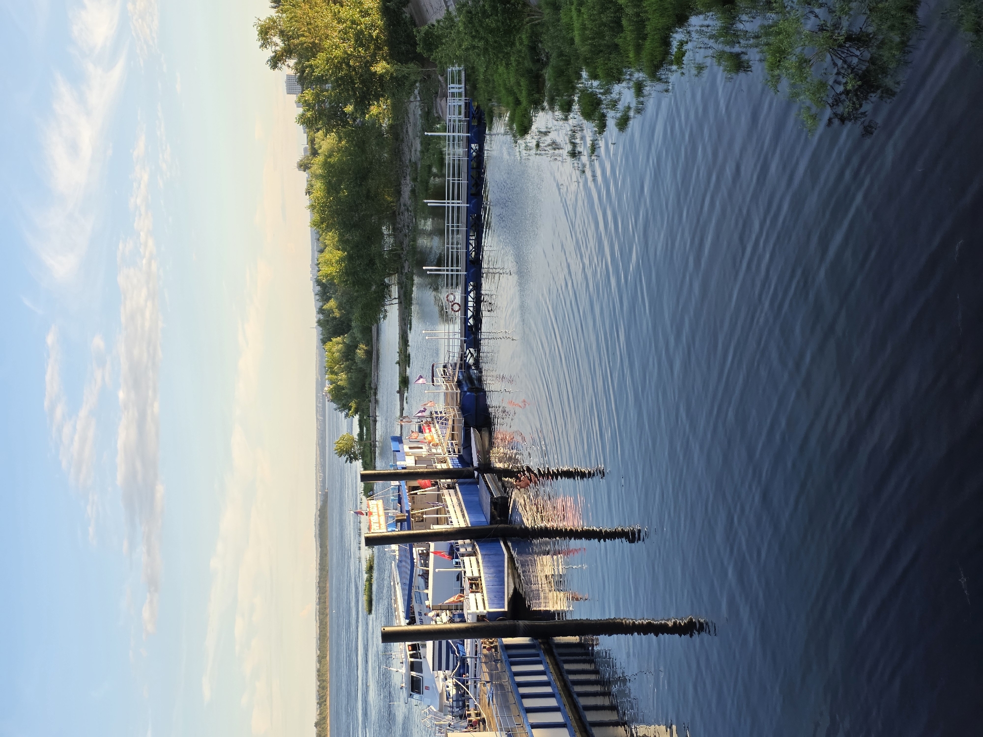 The image displays a body of water with several boats docked alongside a pier. The scene is set along a riverbank lined with lush green trees. The sky is a soft blue with wispy white clouds, suggesting it's either early morning or late afternoon. The water is calm with gentle ripples, reflecting the surrounding trees and the sky.
Several boats are visible, all appearing to be passenger vessels, possibly for tours or transportation. They are docked parallel to the pier, which is a long, elevated structure with railings. The boats are multi-decked and painted in shades of white and blue, with some red accents. Flags are visible on some of the boats.
The pier itself is a prominent feature, with white railings and dark vertical posts. Life rings are also attached to the pier. The reflection of the boats and the pier on the water adds depth to the scene.
The overall atmosphere is peaceful and serene, with a sense of calm that often accompanies riverside settings at this time of day. The location appears to be Volgograd, Russia, as suggested by the context provided. No text is visible in the image.
