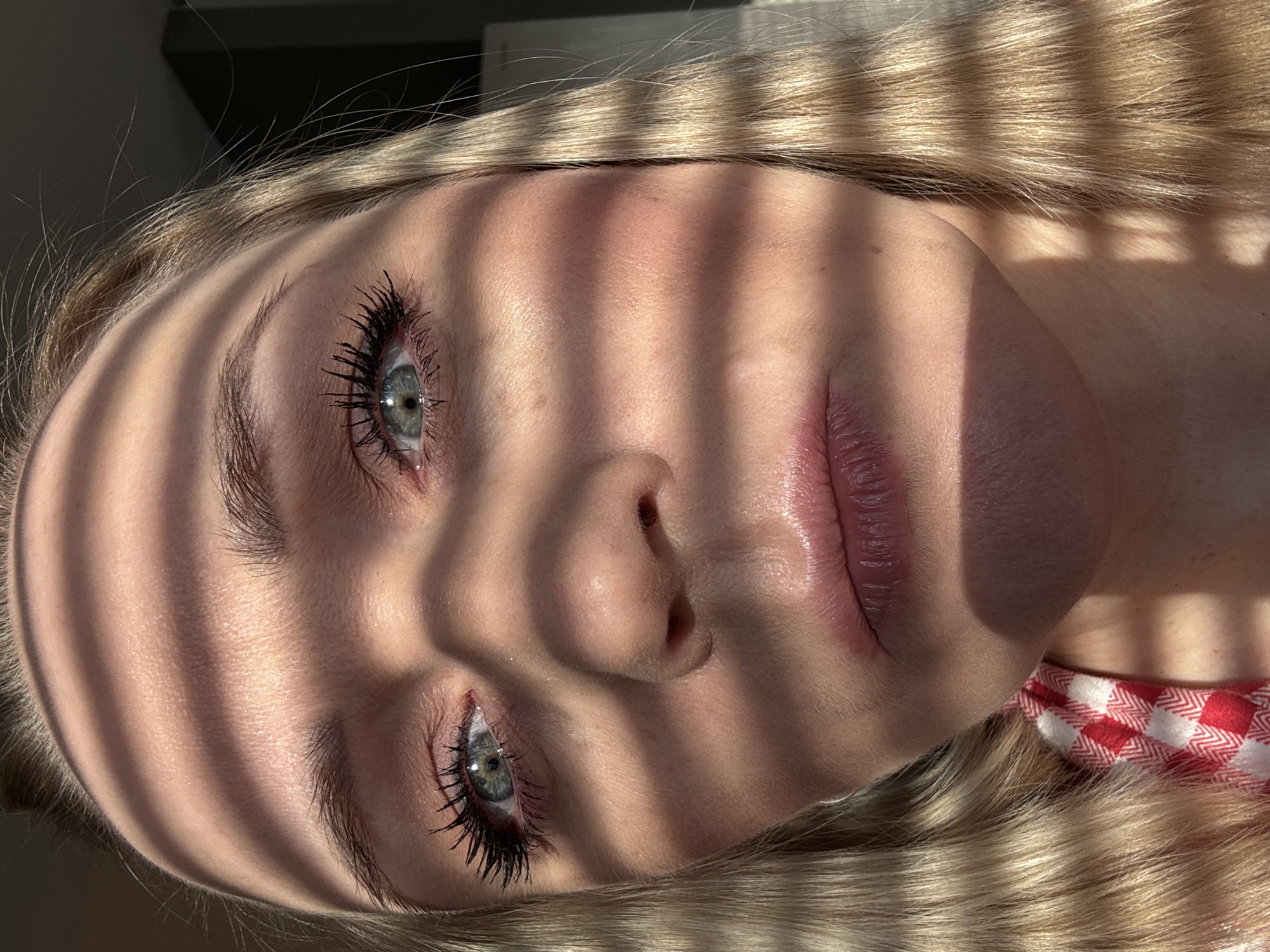A close-up, rotated view shows a person's face partially obscured by shadows cast by parallel blinds or slats. The person has light skin, blonde hair, and blue-green eyes with heavy mascara. Part of a red and white checkered garment is visible at the bottom right corner. The location is not specified, nor is there information to indicate that the location is in the city of Votkinsk, Russia.
