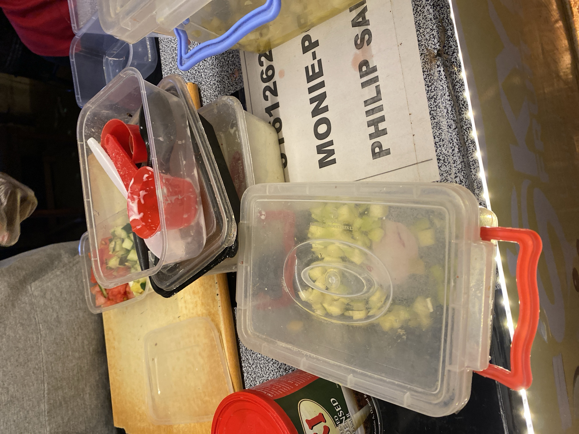 The image shows a collection of plastic food containers on a countertop. Some containers are transparent and contain food, including a diced green vegetable and a tomato-based salad with red and white utensils. There is also a red can with a green label partially visible. In the background, there is a white sign with the text "01262 MONIE-P PHILIP SAL." There is no indication that this is located in Durimi, Nigeria.