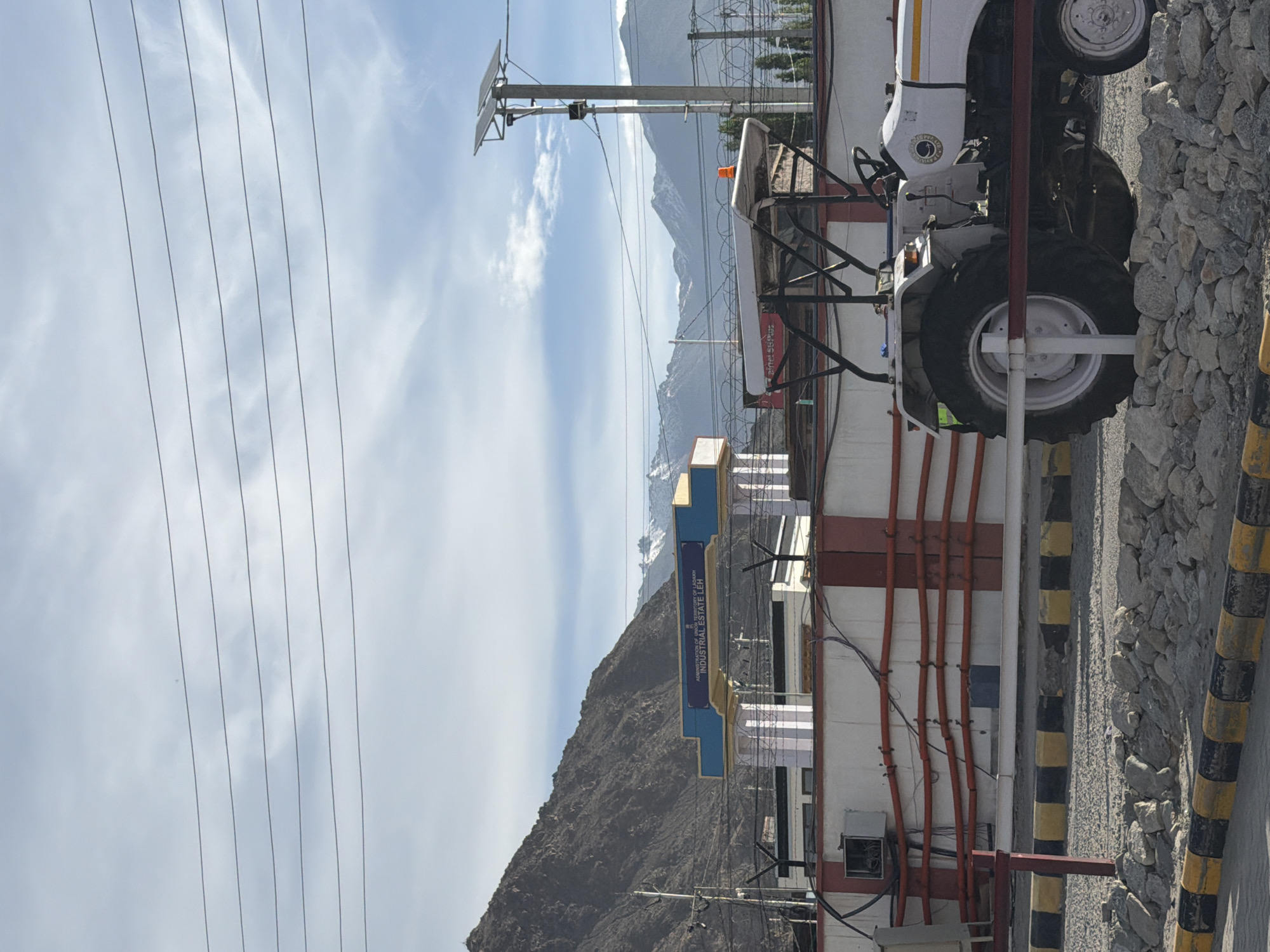 The image shows the "Industrial Estate Leh" archway in Leh, India. The archway is blue and yellow and has text written on it. Behind the archway, a large, rocky mountain range can be seen. In front of the archway, a white tractor is parked. The tractor has a roll cage and an orange flashing light on top. There are also power lines and a solar panel on a pole in the scene. The ground is covered with gravel and rocks. The sky is blue with some clouds.