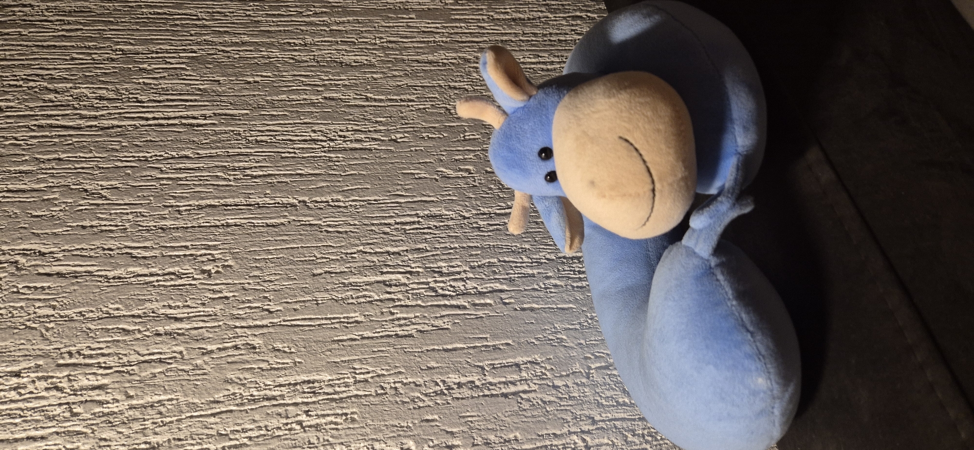 The image shows a blue plush toy animal with a tan face lying on a textured, light-colored surface. The toy is positioned on the right side of the frame, with the left side mostly occupied by the textured surface. A dark area is visible on the far right edge.