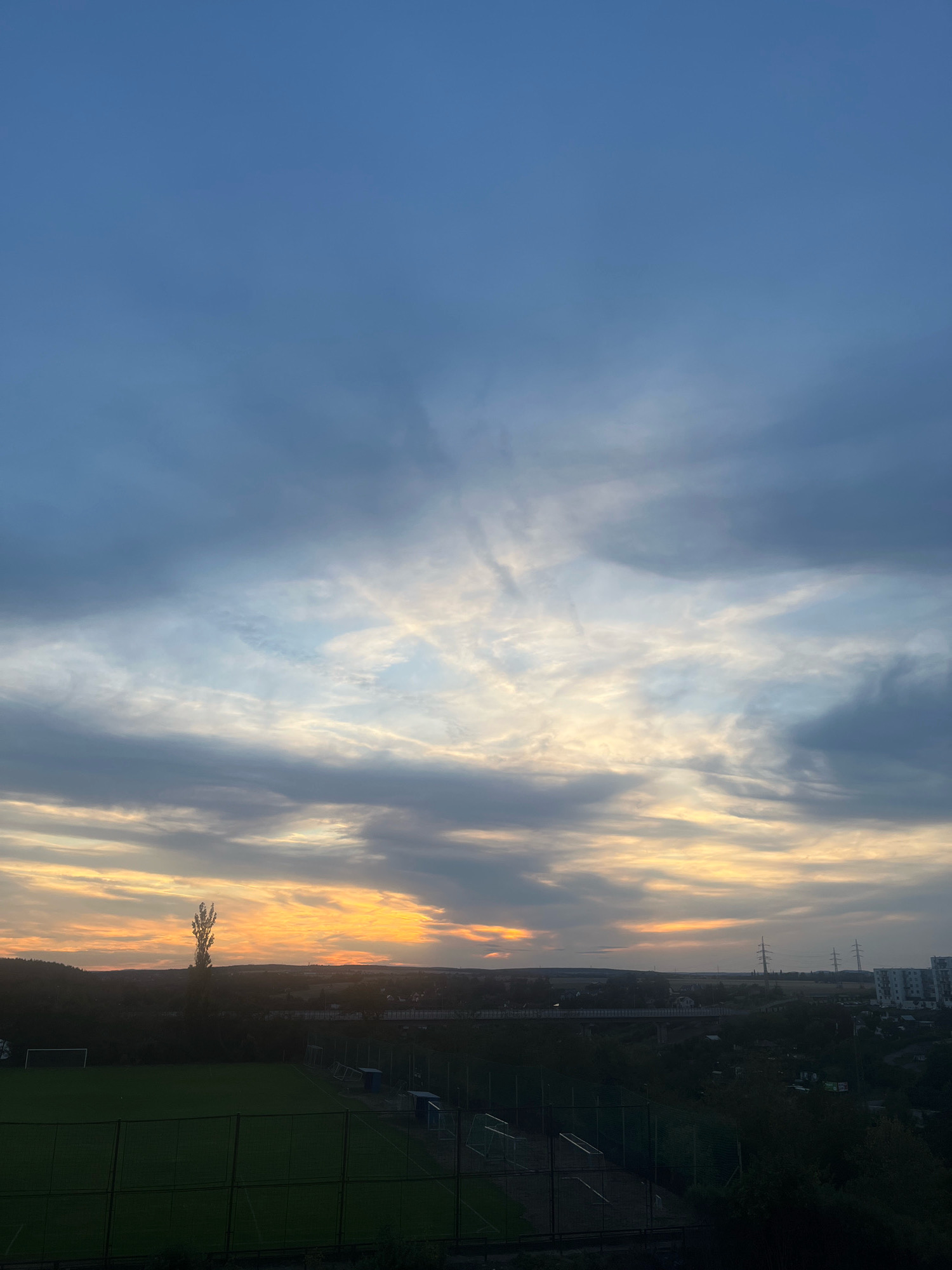 A vertical, medium shot captures a dramatic sunset over a city. The sky is filled with a spectrum of colors, transitioning from a deep blue at the top to vibrant oranges and yellows near the horizon, illuminated by streaks of white and gray clouds. Below the sky, a silhouetted landscape unfolds. A sports field with a net fence is visible in the foreground. Beyond the field, trees and buildings are dimly lit. A tall, slender cell tower stands out against the sunset. The background features a cityscape with apartment buildings and power lines, suggesting an urban environment in Pilsen, Czechia. The overall impression is one of natural beauty contrasting with urban development at dusk.