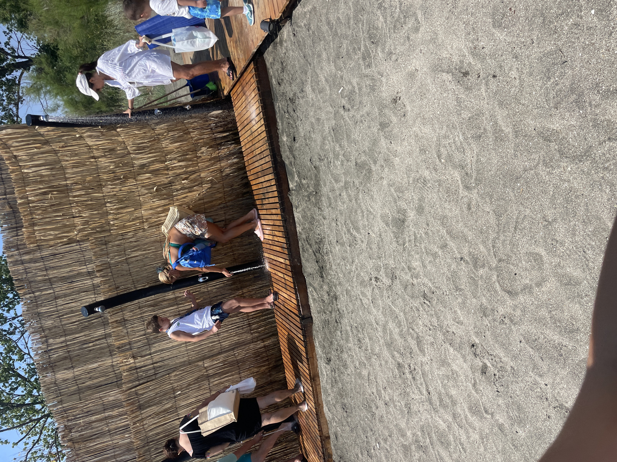 An outdoor shower facility on a sandy beach in Burgas, Bulgaria, is depicted. Several individuals are present, including a woman in a white shirt and hat rinsing under the shower, a boy in a white tank top and shorts standing nearby, and another child in blue shorts. A woman in a black dress with a tote bag is also visible on the left side of the frame, and a person in a green bikini top and patterned shorts is using the shower. The shower structure is made of bamboo reeds, and a wooden platform surrounds the showering area. The ground is covered in sand.