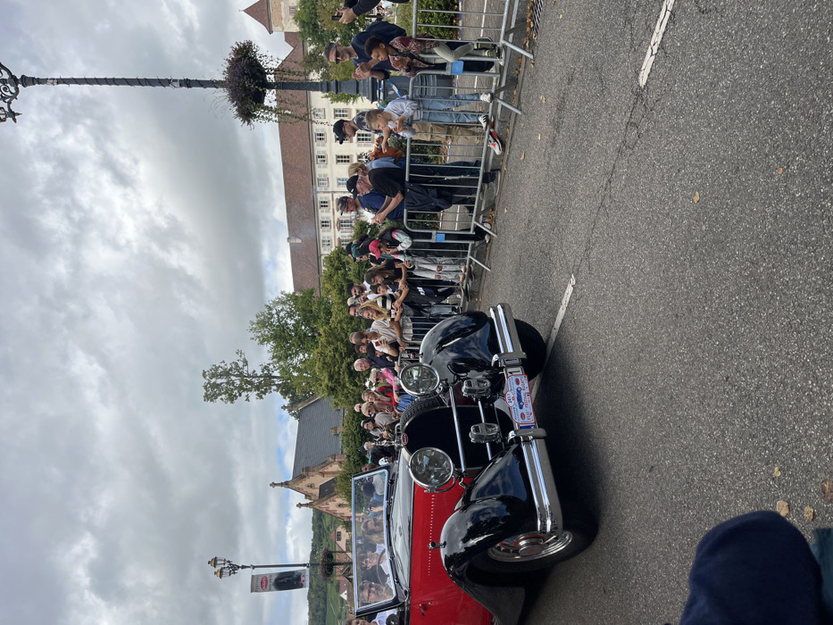The image depicts a public event, likely the Bugatti Festival, in Molsheim, France. A vintage two-tone convertible automobile, predominantly black with a red body section, moves from left to right on an asphalt road. The car features prominent chrome components, including large circular headlights and bumpers, and displays a special event placard on its front bumper identifying it with "BUGATTI FESTIVAL MOLSHEIM" and the number "1." Multiple occupants, including a male driver wearing glasses, are visible within the vehicle.
On the right side of the road, a crowd of spectators of varying ages, including adults and children, is gathered behind silver metal crowd control barriers. Many individuals in the crowd are holding smartphones to record the event, and some adults are seen clapping. Children in the front row are observed standing on the lower bar of the barriers.
The background reveals several distinct architectural features of Molsheim. To the left-midground, an ornate, gothic-style stone chapel with a reddish-brown tiled roof is visible, partially obscured by green foliage. This building strongly resembles the Chartreuse de Molsheim. To the right-midground, a multi-story building with a reddish-brown tiled roof and light-colored facade is present. Further to the right, a structure featuring a turret-like element is visible, indicative of historic town architecture. Distant vineyards are visible on a hillside to the far left. The scene is illuminated by diffused natural light under an overcast sky. Two ornate black lampposts are present, one on the left displaying a banner that reads "BUGATTI FESTIVAL MOLSHEIM," and another on the right adorned with a hanging plant basket.