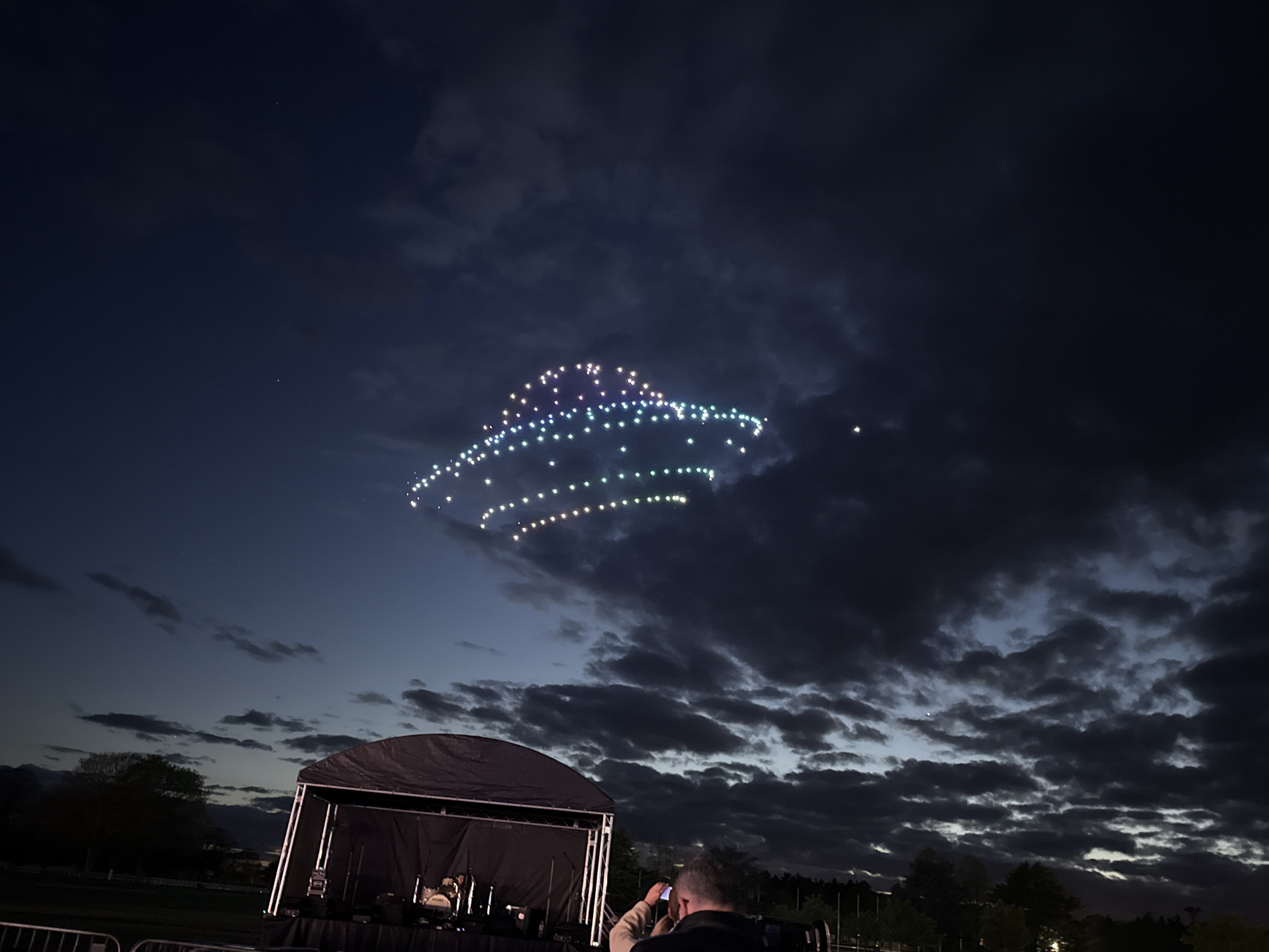 A nighttime scene in South Norfolk, United Kingdom, showing a stage with equipment in the foreground. Behind the stage, a person holds up a mobile phone, potentially photographing or recording the scene. The sky is dark with heavy clouds. In the upper part of the frame, an illuminated aerial display of lights in the shape of a rounded object is suspended against the night sky. Trees are visible in the background.