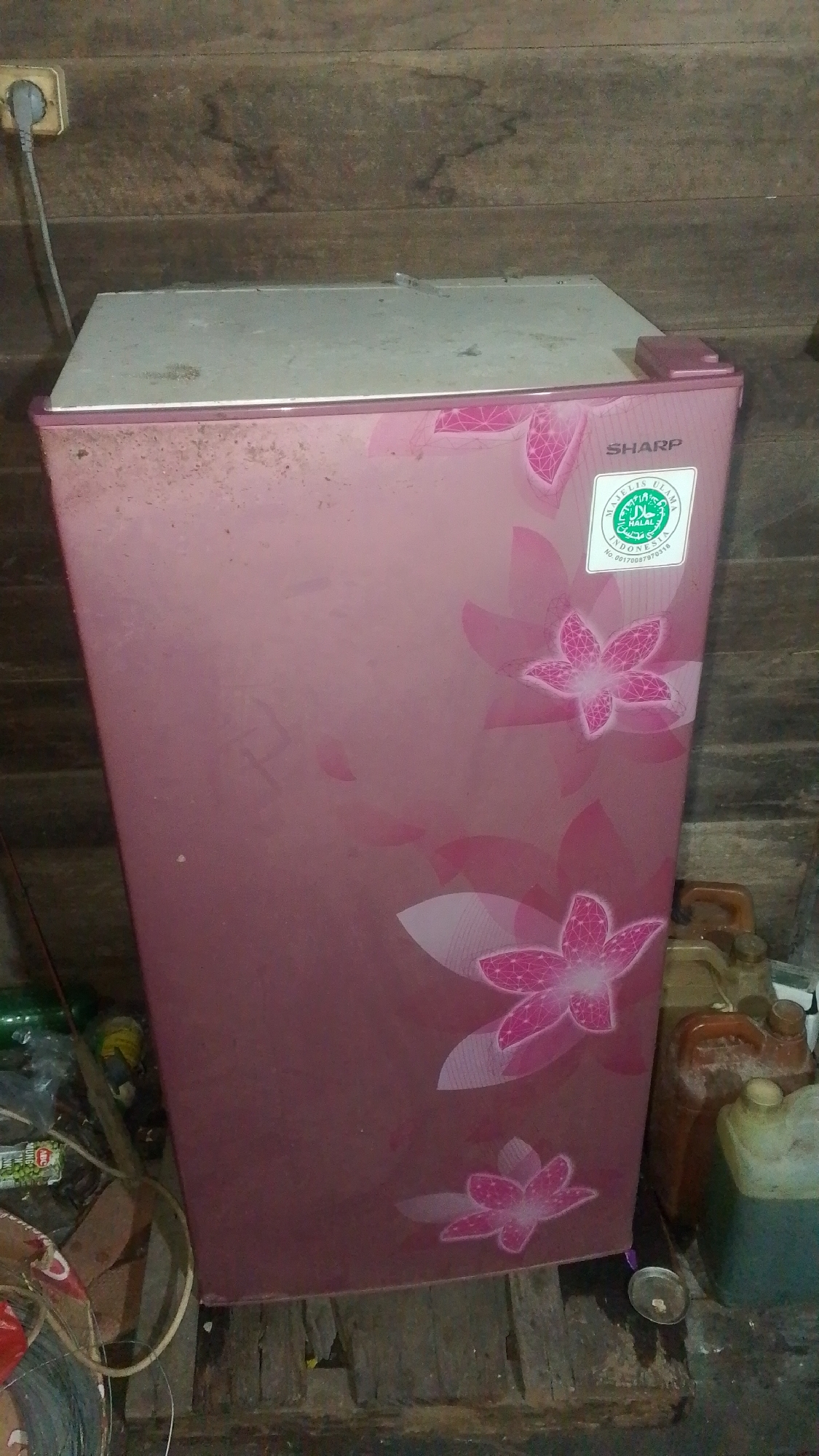 A pink refrigerator with a floral pattern is positioned in the center of the frame. It is standing on a wooden floor. The top of the refrigerator appears to be a light gray color and is dusty. On the right side of the refrigerator door, there is a circular green and white sticker with writing on it. To the right of the refrigerator, there are several plastic containers, some of which appear to be jerry cans. In the background, there is a wooden wall. To the left of the refrigerator, there are some items on the floor, including plastic bags and a yellow cylindrical object. A white electrical cord is plugged into an outlet on the wooden wall above the refrigerator. The overall environment appears to be utilitarian, possibly a storage area or a less-maintained part of a dwelling.