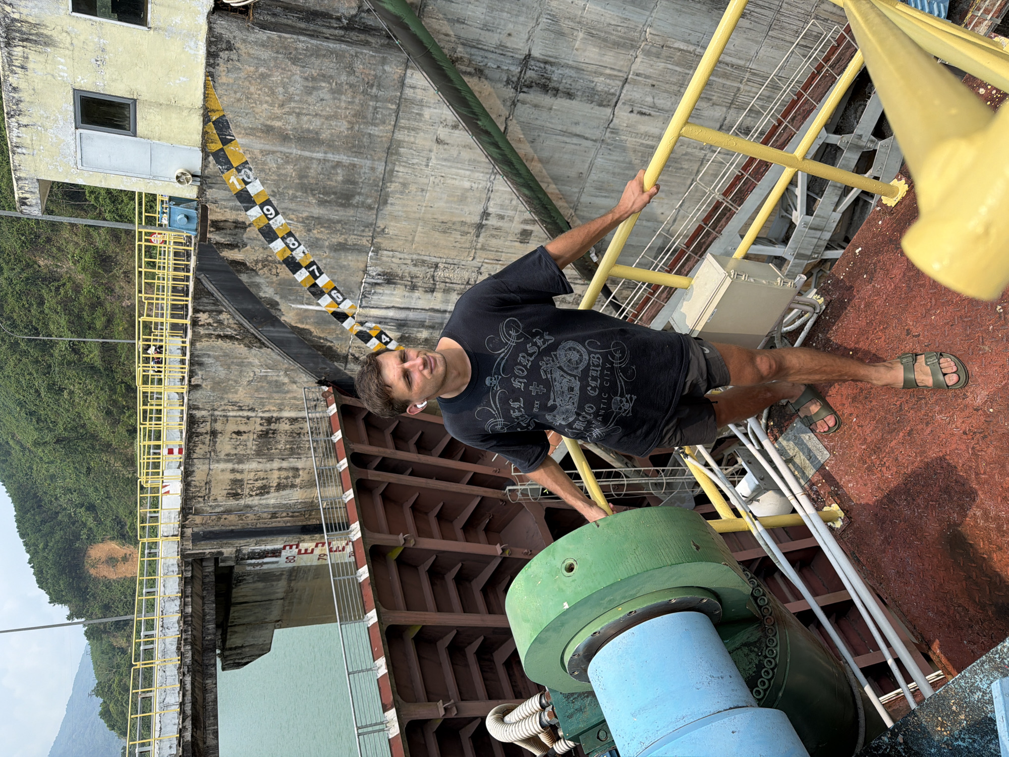 A man is standing on a painted-red surface of what appears to be a dock, clinging to yellow handrails. He is wearing a dark T-shirt, dark shorts, and sandals. Behind him is a concrete dam structure with a water level marker. There are metal and structural elements visible, including a large green and blue cylindrical machine. In the background, there is a lush green landscape and the sky. Based on the location clues, it's plausible that this scene is near a hydroelectric power plant in Đà Nẵng, Vietnam.