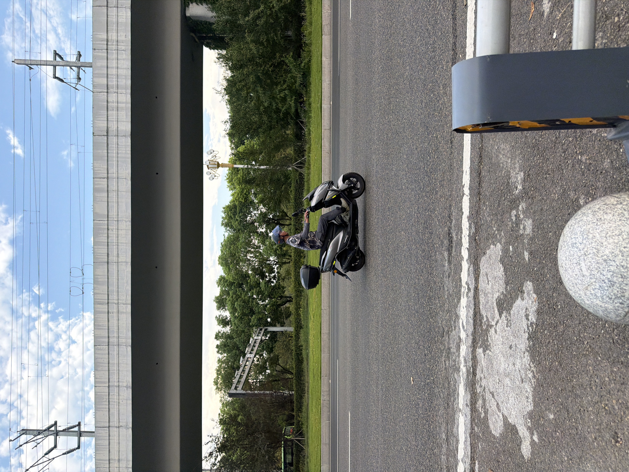 A person wearing a helmet rides a grey scooter on a paved road. The scooter has a black storage box on the back. The person is wearing a patterned shirt and dark pants. In the background, there is a large concrete overpass with power lines running overhead. Trees and green foliage line the side of the road. A street lamp is visible in the distance. The foreground shows a textured grey surface, possibly part of a barrier, and a portion of a grey, speckled spherical object. The scene appears to be an urban environment in Jiangjin, China.