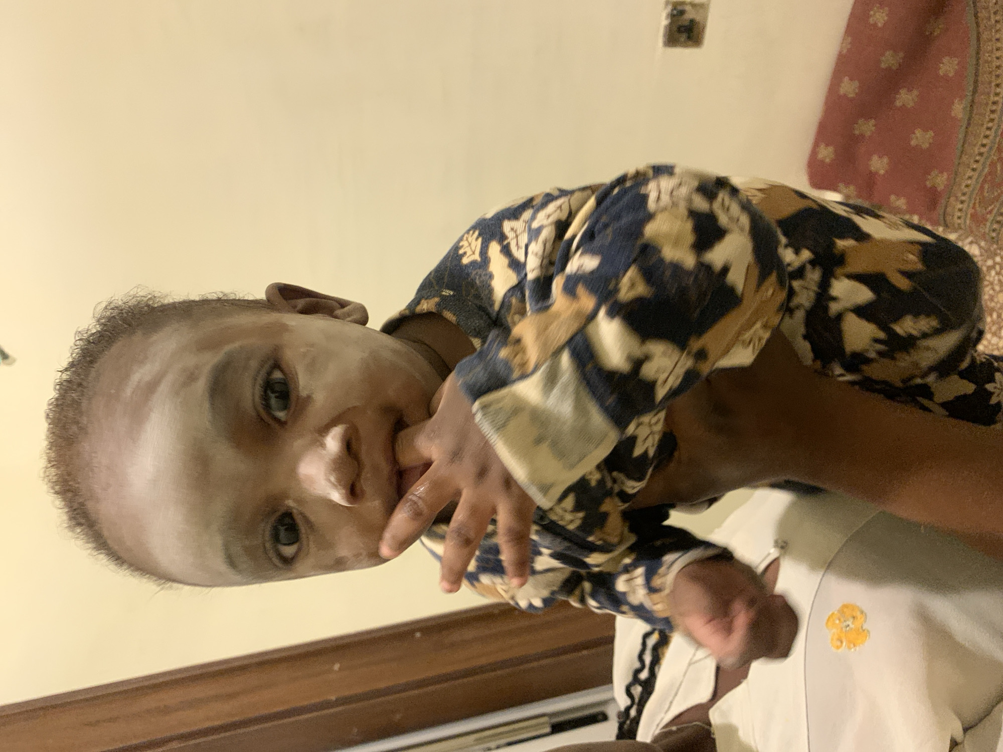 A close-up shot depicts a baby being held by an adult indoors, possibly in a residence in Jos, Nigeria. The baby has a grey-toned face and is wearing a patterned long-sleeved shirt with blue, brown, and beige tones. The baby's fingers are near their mouth. An adult's arm is visible supporting the baby. Partially visible is another baby in a white garment bearing a yellow teddy bear emblem. Behind the subject, there is a wall with a visible electrical outlet, along with a portion of a decorative red fabric hanging. A wooden doorframe is visible at the bottom of the frame. The image is oriented vertically.