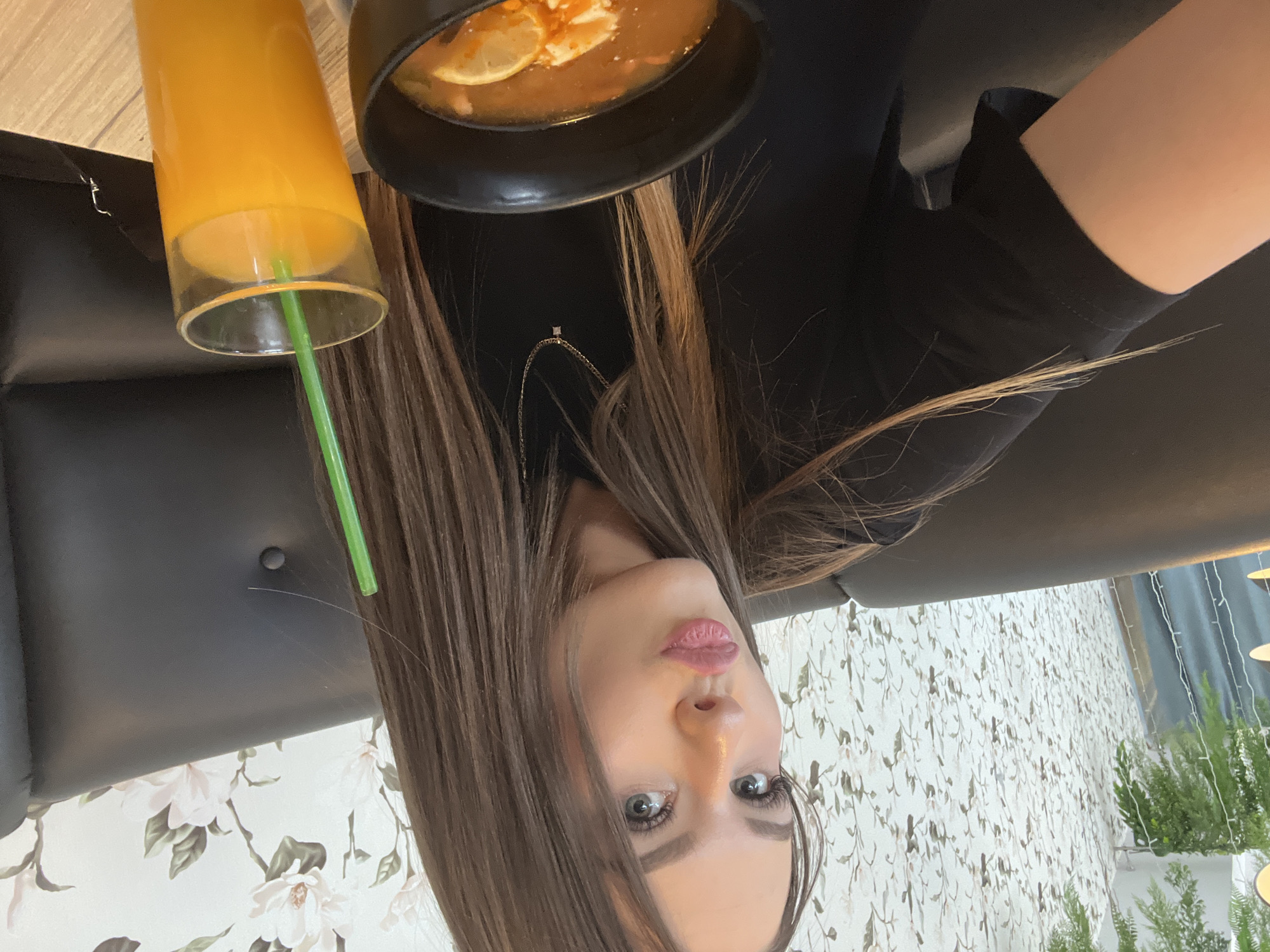 The image shows a woman with long brown hair in a restaurant. She is wearing a black top. A glass of orange juice with a green straw and a black bowl containing soup with lemon slices are on the table. The restaurant has floral wallpaper and plants. The location is Chaikovsky, Russia.