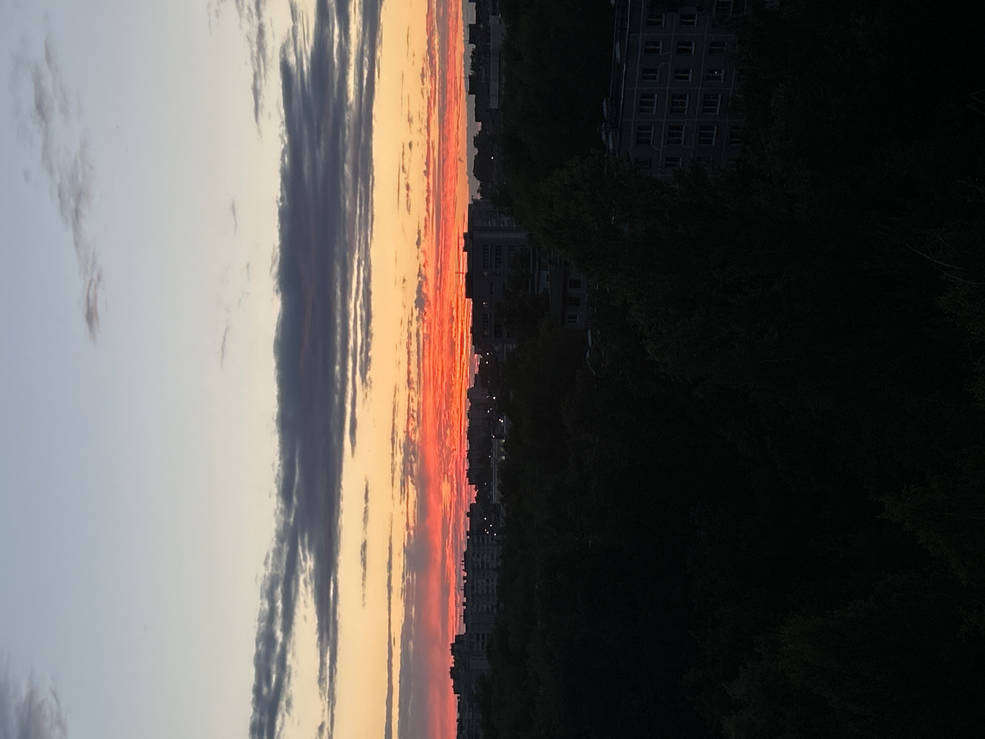 This is a full shot of a city skyline at dusk, with the sun low on the horizon. The sky is a gradient of soft blue at the top, transitioning to hues of orange and fiery red as it nears the horizon. A band of dark, wispy clouds stretches across the middle of the sky. Below the sky, silhouetted trees and buildings form the urban landscape. The buildings are predominantly multi-story apartment blocks, some with visible windows. The foreground is filled with the dark mass of trees. The lighting suggests it is either sunrise or sunset.