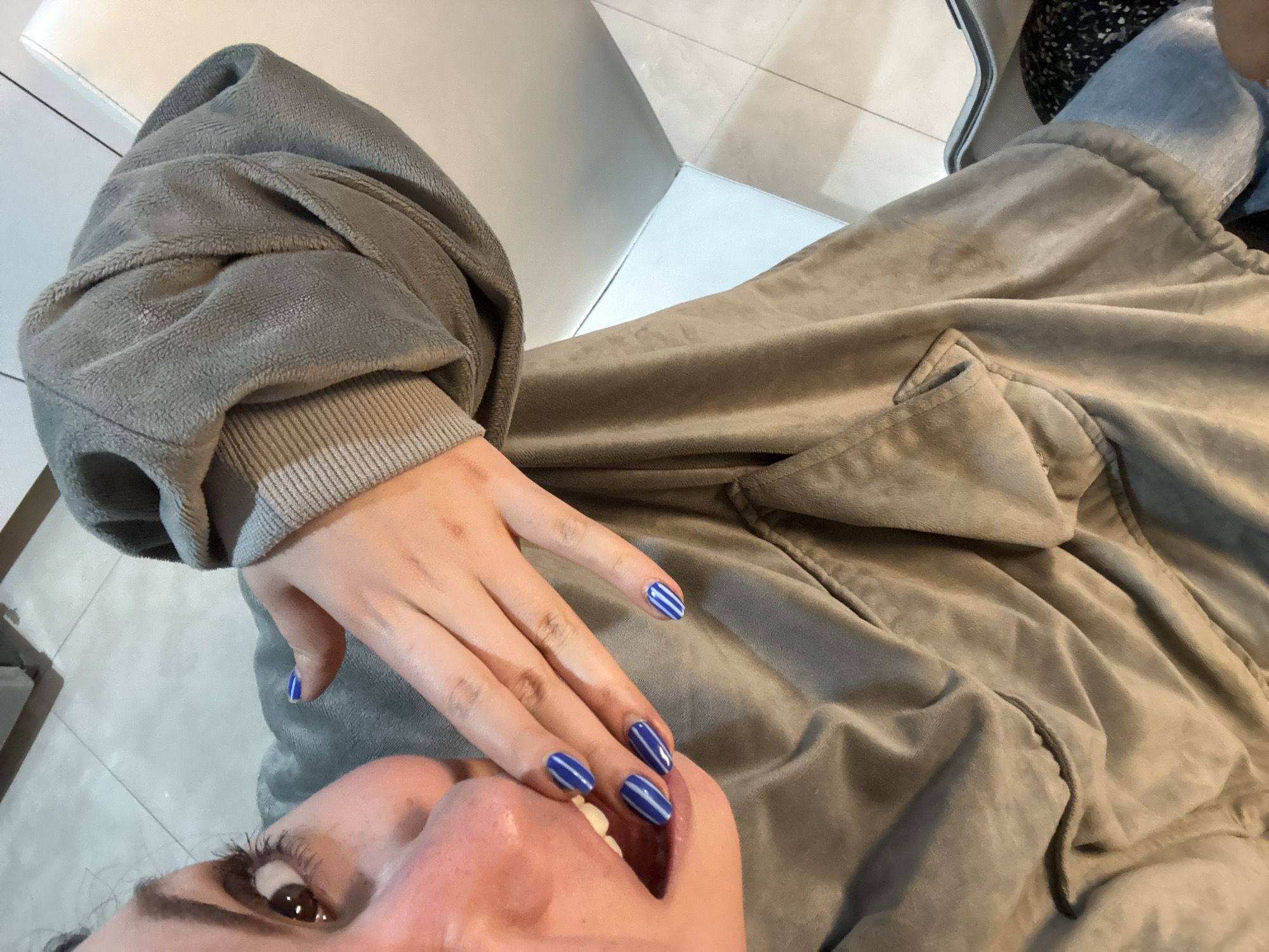 A close-up, high-angle shot shows a person wearing a grey, plush jacket, appearing to be indoors. The person's face is partially visible in the lower part of the frame. The person's hand rests on their face, displaying blue nail polish with white stripes. The jacket is textured. The background is a mix of white surfaces, appearing to be walls and possibly flooring. The location, Santa Clara Ocoyucan, Mexico, cannot be confirmed from the visual information. There is nothing to give a location clue.