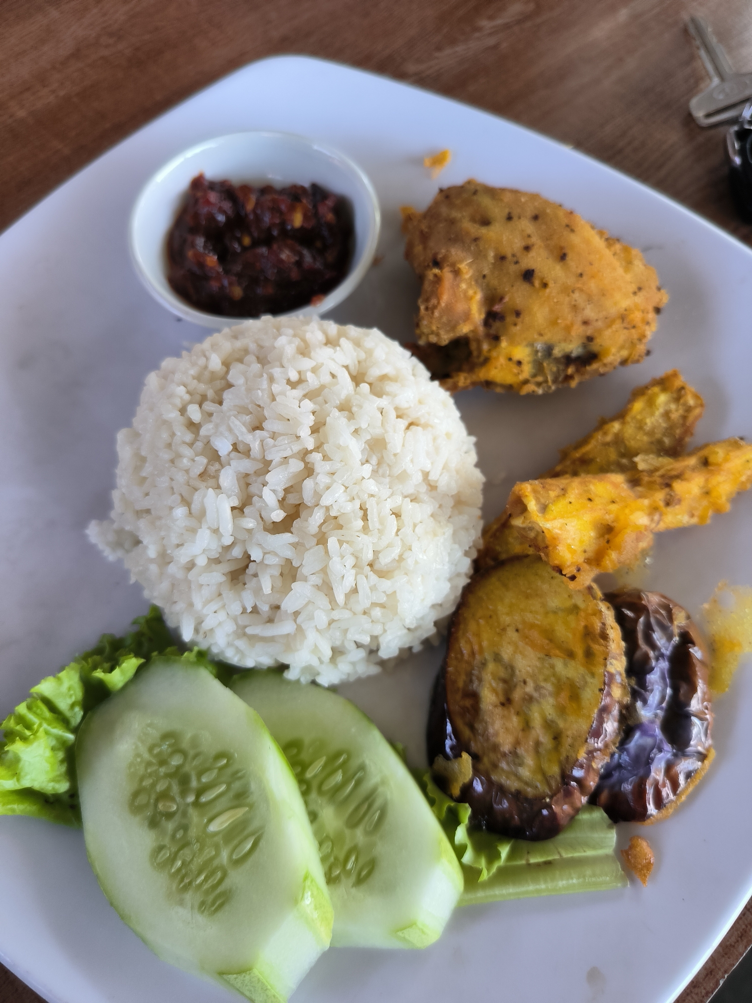 On a square white plate, a meal is arranged. A mound of white rice is centrally located. To the left, two cucumber slices lie on a bed of lettuce. Adjacent to the rice, a small white bowl contains a dark red sauce. To the right of the rice, there's a piece of fried chicken along with fried bananas, and fried eggplant. This scene likely takes place in Matang, Indonesia. The presence of a car key at the top-right corner suggests the plate is on a table, possibly in a restaurant or home setting.