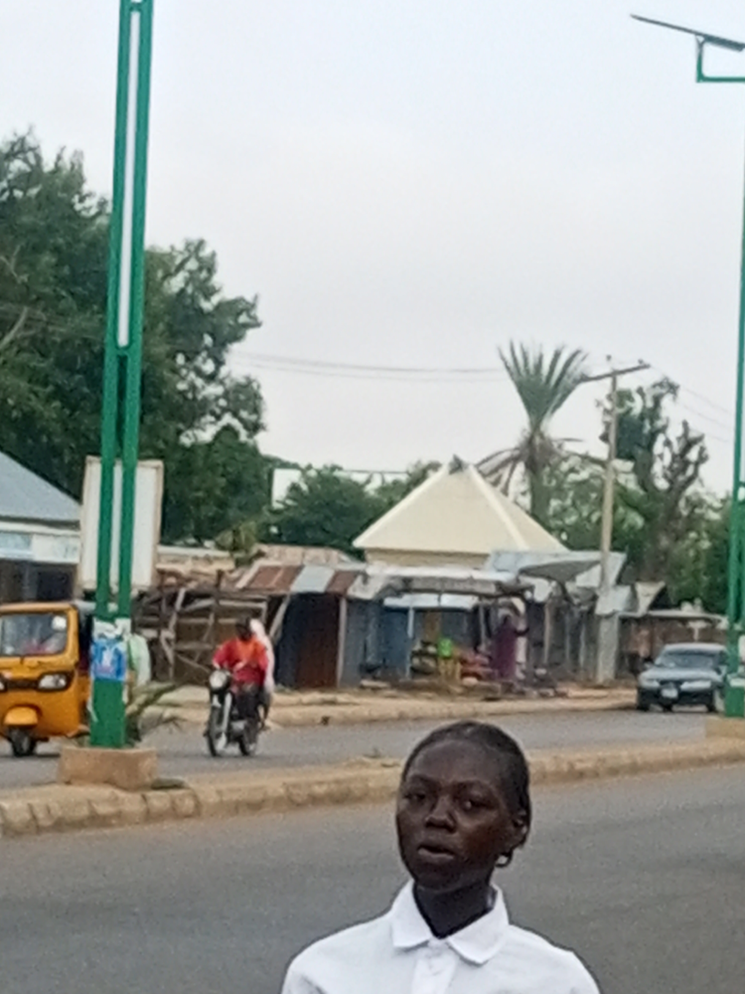 In Garko, Nigeria, an outdoor scene captured on an overcast day shows a young woman in the foreground, wearing a white collared shirt. Behind her, across a paved road, is a marketplace with several stalls. A yellow auto-rickshaw is parked to the left of the marketplace. To the right of the auto-rickshaw, a person in an orange shirt rides a motorcycle. In the background, there are trees and power lines. A silver car is visible further down the road to the right.