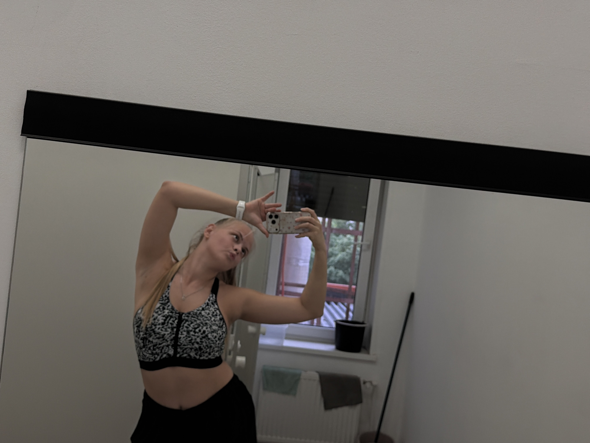 A woman is taking a selfie in front of a mirror. The mirror has a black frame. The woman has blonde hair pulled back in a ponytail and is wearing a black and white patterned sports bra and black bottoms. She is holding a smartphone in her right hand, presumably to take the photo. Her left arm is raised above her head. Behind her, the mirror reflects a room with a window overlooking what appears to be a residential area. There is a radiator under the window and a cleaning stick to the right. The location details, specifically Izhevsk, Russia, cannot be verified with the information available in this image.