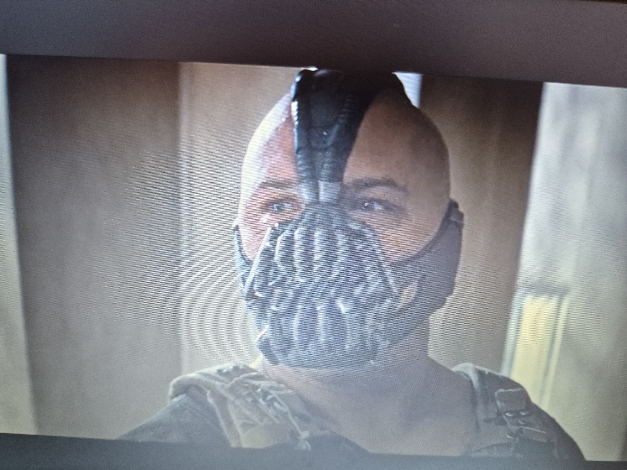 The image shows a close-up of a bald man wearing a dark, tactical mask that covers his nose and mouth. The mask has a ribbed texture around the mouth area and appears to be connected to a headpiece that extends upwards. The man's eyes are visible, and he has a serious expression. He is wearing a dark, possibly military-style outfit with straps and buckles across his chest. The background is blurred and appears to be an indoor setting with muted, earthy tones. There is no indication of the city of Sandwell, United Kingdom, or any specific geographical location.