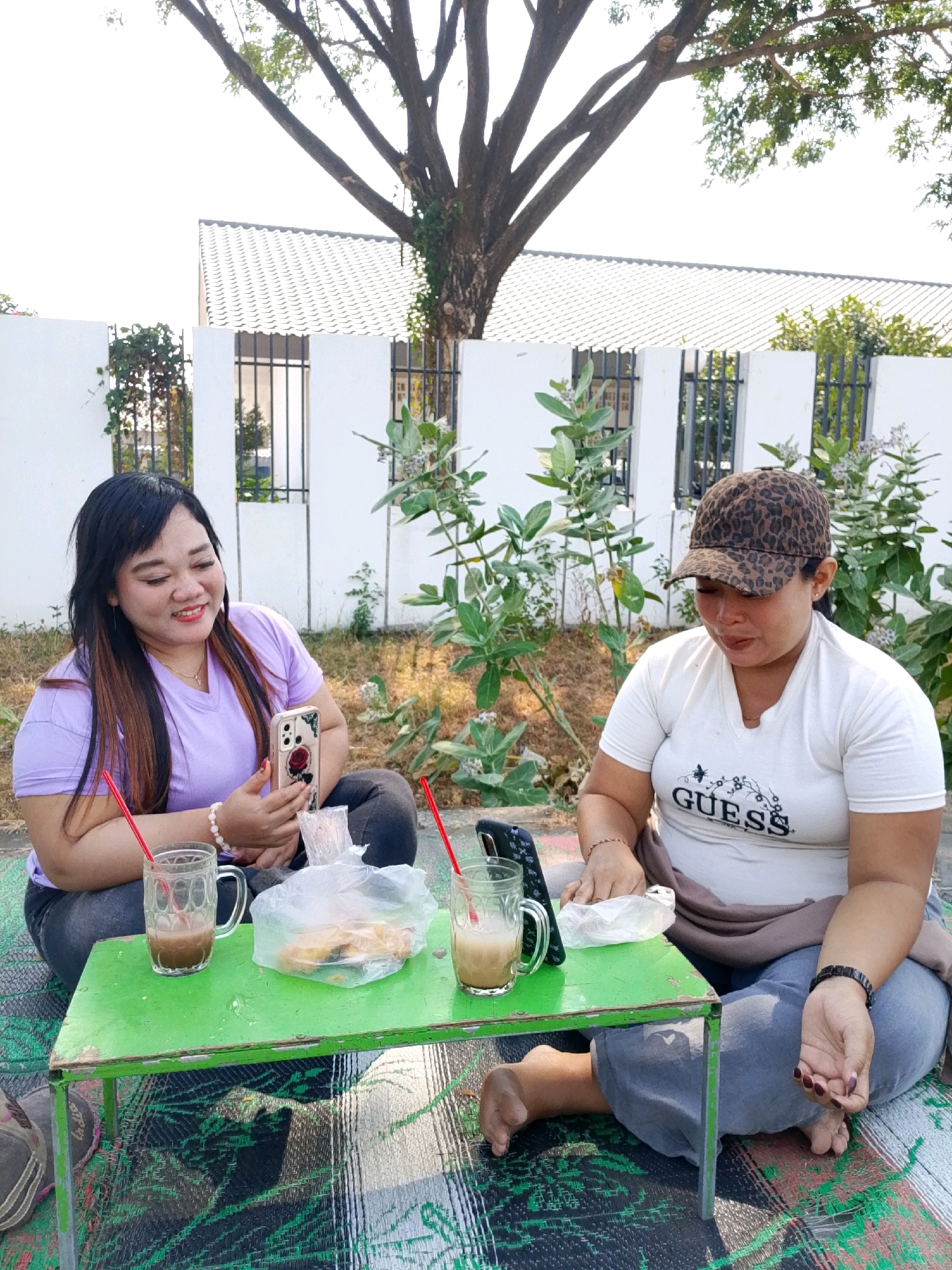 Two women are seated on a patterned mat outdoors in Bulusari, Indonesia, facing a small green table. The woman on the left, wearing a lavender t-shirt, is holding a smartphone with a rose design case and looking at it. She has long, dark hair and is wearing a white beaded bracelet. The woman on the right is wearing a white t-shirt with the word "GUESS" printed on it and a leopard-print baseball cap. She is looking down at the table. On the table, there are two glasses with red straws and a beverage, a plastic bag containing food items, and a second smartphone. Behind the women, a white building with a tiled roof and barred windows is visible, along with some green foliage and a large tree. The ground appears to be dry grass and dirt. The overall scene suggests a casual outdoor gathering.