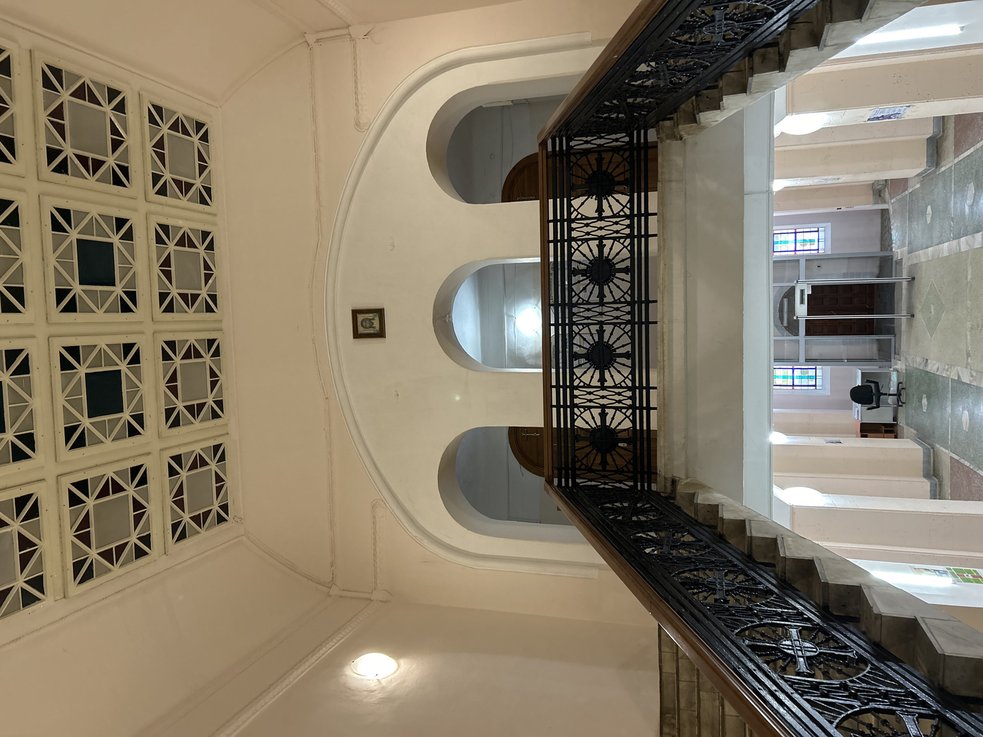 The image shows a wide staircase with ornate black metal railings and dark wood handrails, ascending towards a landing. Above the landing, there are three arched doorways. A small framed picture of Jesus Christ is visible on the wall above the central arch. The ceiling features a decorative grid pattern with square and triangular colored inlays. Downstairs, there is a security gate, a desk with a chair, and stained-glass windows. The building appears to be a public space, possibly a church or a historical building. This location is the interior of the Tomsk Spiritual Seminary in Tomsk, Russia.