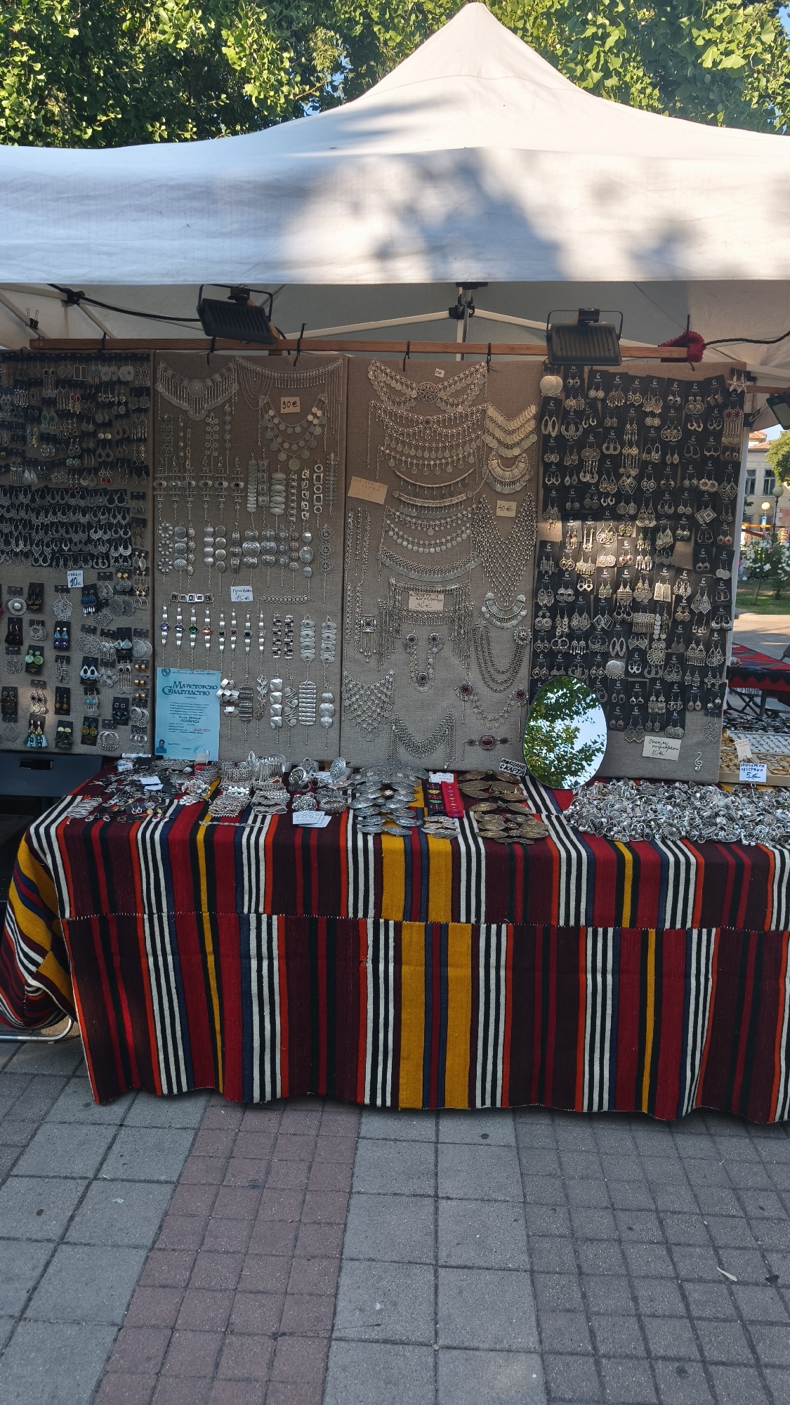 This is an outdoor market stall in Pomorie, Bulgaria, featuring an assortment of silver-colored jewelry displayed on fabric-covered boards. The jewelry includes necklaces, earrings, and bracelets. The stall is covered by a white tent, and the display table is draped with a colorful, striped cloth. The ground is paved with cobblestones. A small, oval mirror is propped up on the table, reflecting the trees and sky. There are no people clearly visible in the image.