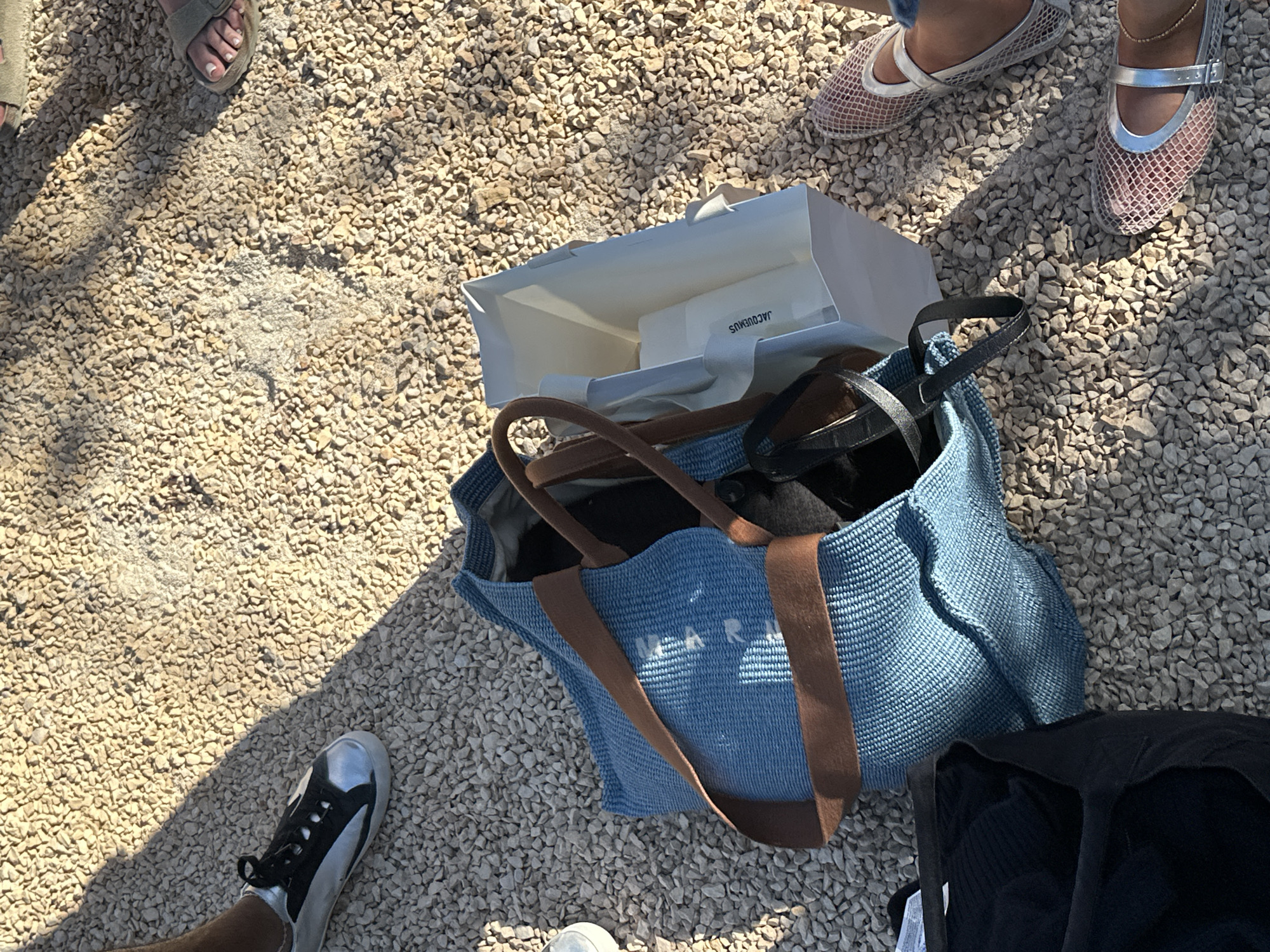 Captured from an overhead perspective, the image depicts a scene on a gravelly ground, likely in Sant Josep de sa Talaia, Spain, given its Mediterranean aesthetic. In the composition, one can identify several pairs of feet and footwear: one pair wearing sandals, another wearing mesh ballet flats, and a single foot clad in a black and white sneaker. At the center lies a light blue woven tote bag with brown straps, and a white shopping bag with "JACQUEMUS" printed on it is inside of it. Also near the tote bag is a black cloth. The gravelly ground serves as the backdrop, suggesting an outdoor setting under daylight conditions, evidenced by the presence of shadows.