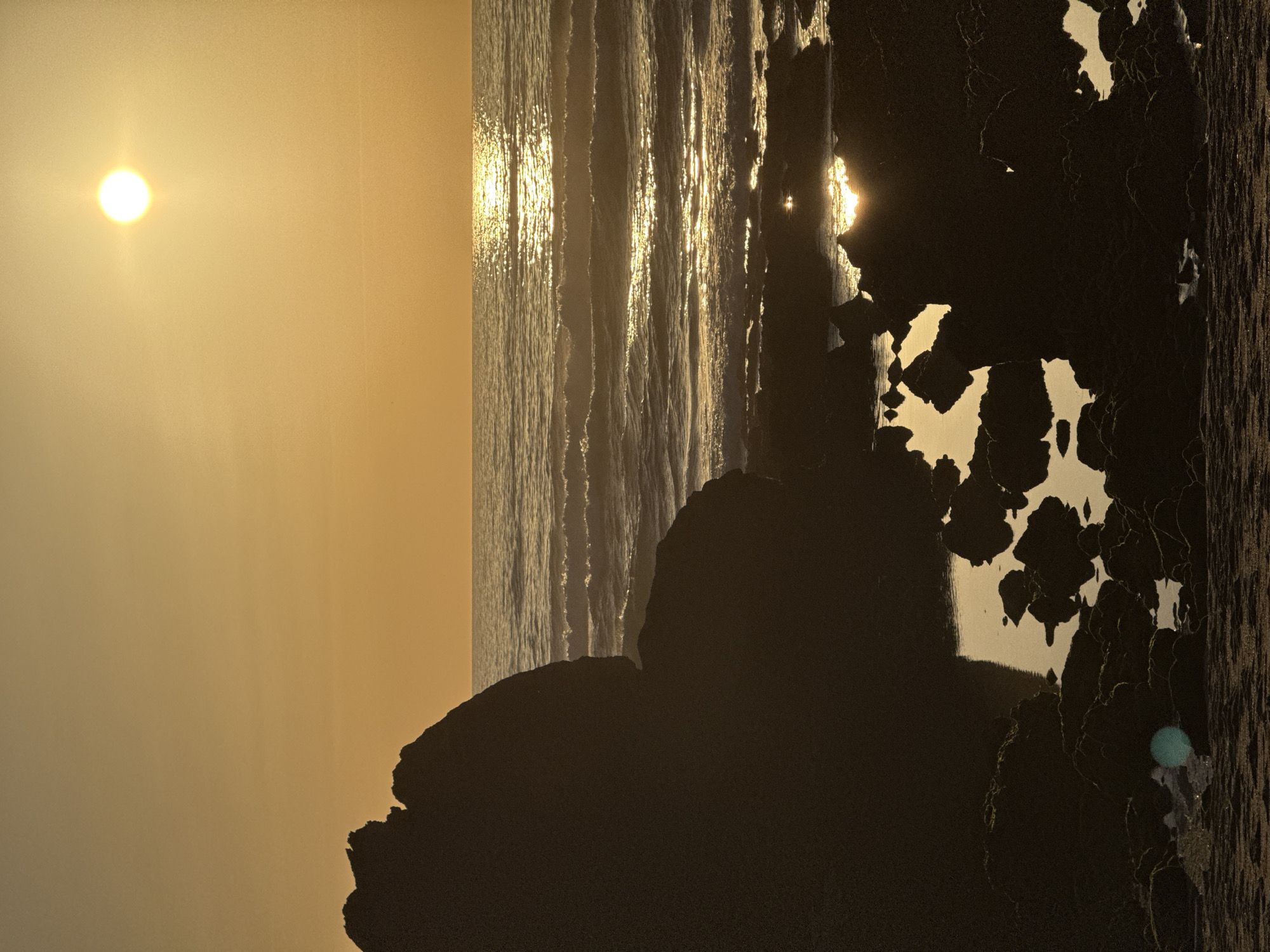 A sunset over the ocean is depicted, with the sun a bright orb in the upper right quadrant of the frame, casting a golden reflection on the water. The waves are gently rolling towards a rocky shore in the foreground. The rocks are dark and silhouetted against the bright water and sky. In the bottom left corner, there is a large, dark rock formation that dominates the left side of the image, its jagged edges outlined by the fading sunlight. The sky is a warm gradient of orange and yellow. The scene appears to be a natural coastal environment, possibly a beach or coastline. The image does not contain any specific landmarks that would definitively place it in Biarritz, France, but it is a generic coastal sunset scene. There are no people visible in the image.