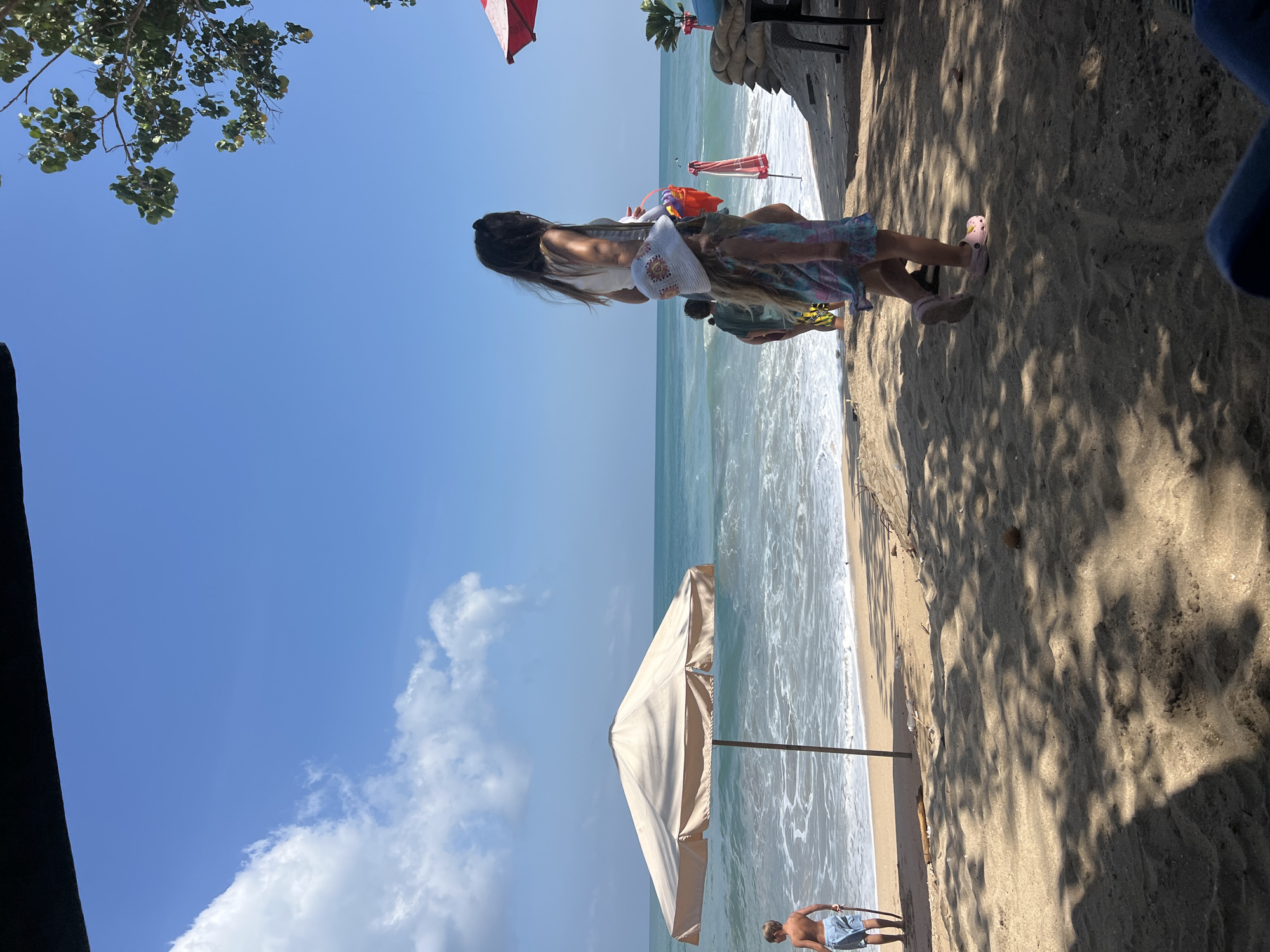 The image shows a beach scene in Kuta, Indonesia. In the foreground, sand is visible with shadows cast by trees. Moving towards the background, there are two young girls and an adult woman walking on the sand, with the ocean and sky in the distance. The woman appears to be holding a bucket. A beach umbrella stands in the middle ground. A young boy in blue shorts is seen walking on the beach near the water. The ocean is calm with gentle waves. The sky is clear and blue with scattered clouds.