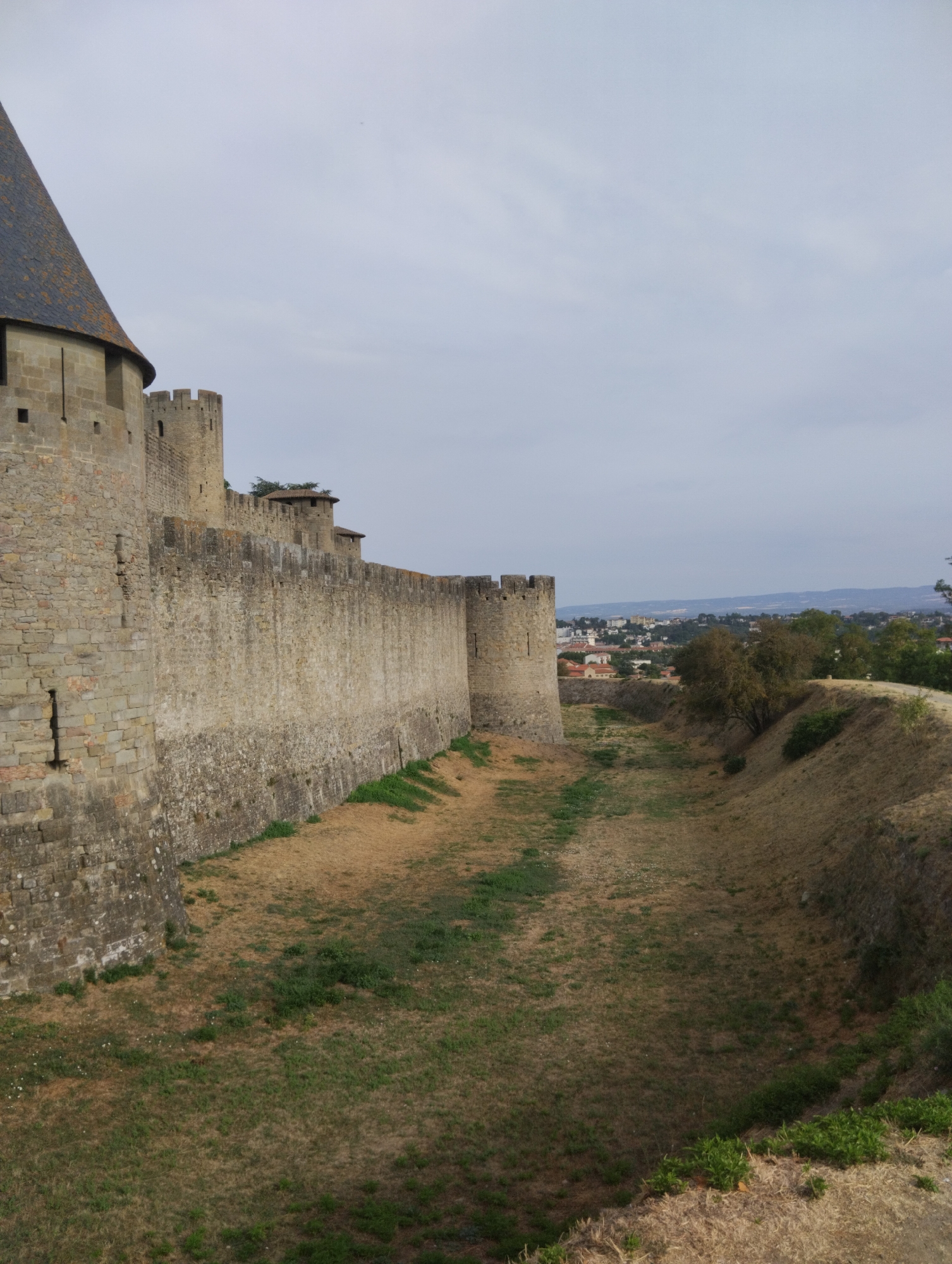 This image shows a view of the fortified city of Carcassonne, France, specifically looking along the outer ramparts and a dry moat. The massive stone walls, constructed from roughly hewn blocks, rise prominently on the left side of the frame. A conical, slate-tiled tower, part of the fortification, is visible in the upper left corner. Further along the wall, crenellated towers and battlements can be seen, interspersed with smaller turrets. The moat, a deep, dry ditch, occupies the right side of the image, its slopes covered with dry, sparse vegetation and some scattered green growth. In the distance, beyond the walls, a cityscape with houses and buildings is visible under an overcast sky. The overall impression is one of historical grandeur and formidable defense.