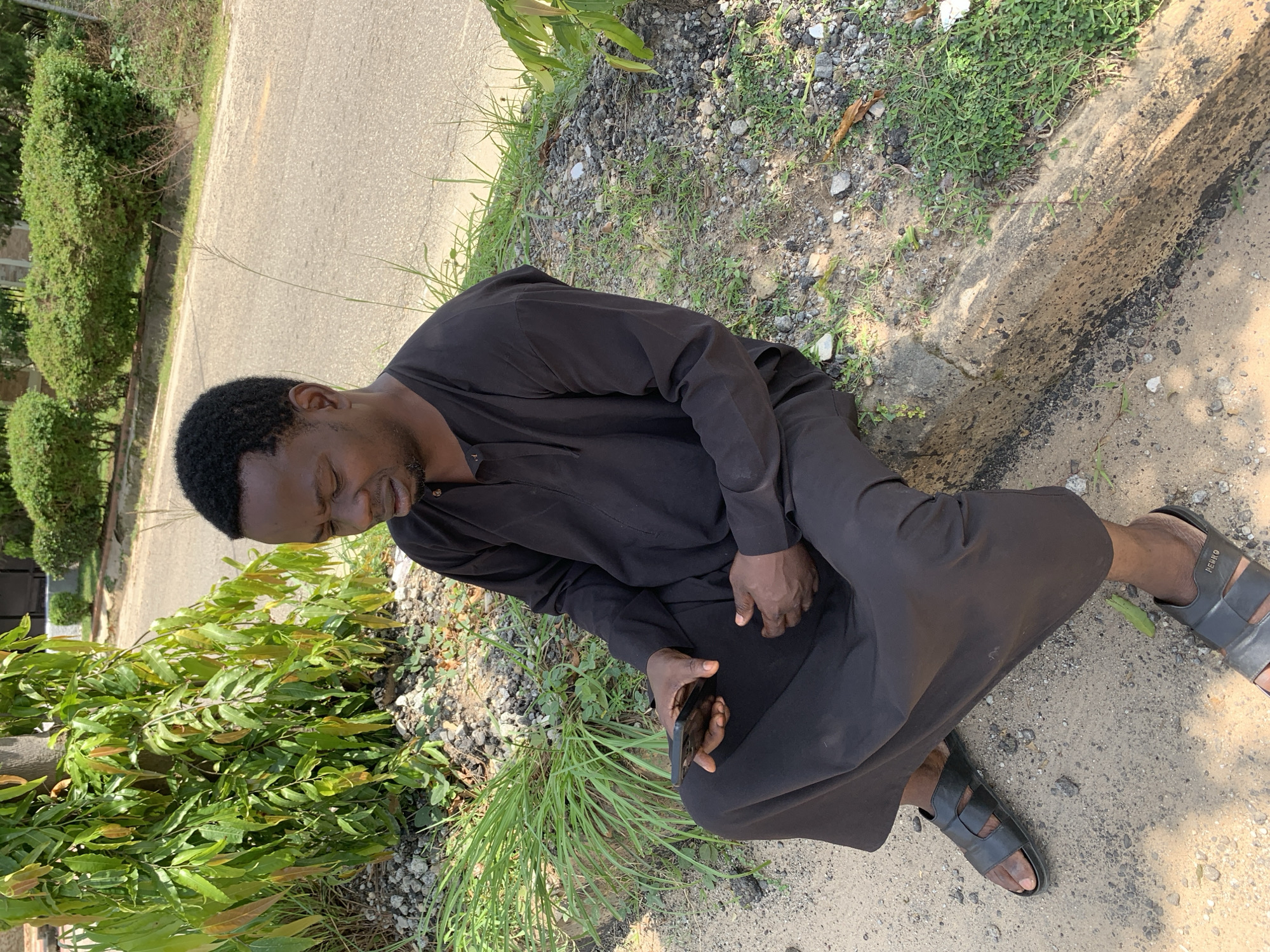 In Abuja, Nigeria, a man is seated on a concrete curb outdoors. He is wearing a black traditional robe and black sandals. He is holding a black mobile phone in his left hand and looking down at it. The background shows a road and some green vegetation. The sun is shining, casting shadows. The ground is a mixture of sand and small stones.