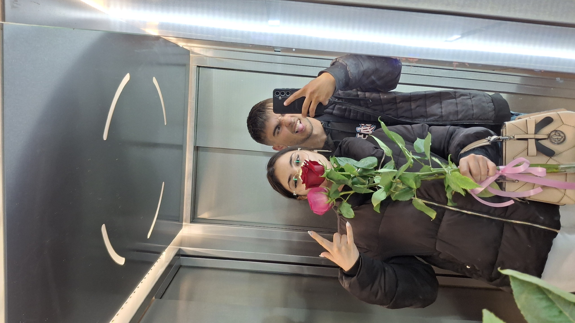 A man and a woman are taking a selfie in an elevator. The man is holding a black smartphone and making a face. The woman is holding a bouquet of roses wrapped with a pink ribbon and making a peace sign with her left hand. She is wearing glasses and a black puffer jacket. A tan purse is adjacent to the bouquet. The elevator features a metallic interior with a pattern of curved lines on one wall. The location is likely in Varna, Bulgaria.