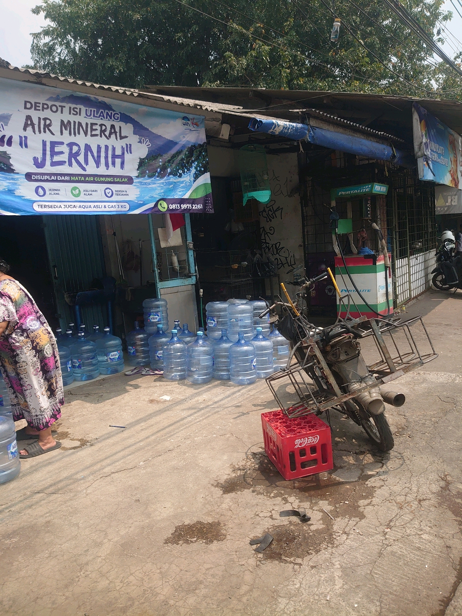A water depot with large blue water jugs is located on a street in Depok, Indonesia. A banner for "AIR MINERAL JERNIH" hangs across the front of the depot. The depot appears to be a small, open-air structure with a corrugated metal roof. In front of the depot, stacks of water jugs are arranged on the ground. A motorcycle with a large metal basket is parked near the water jugs. A red Coca-Cola crate is placed on the ground in front of the motorcycle. The ground is made of concrete and appears wet in some areas. The overall scene suggests a business operating on a street with moderate pedestrian and vehicle traffic.