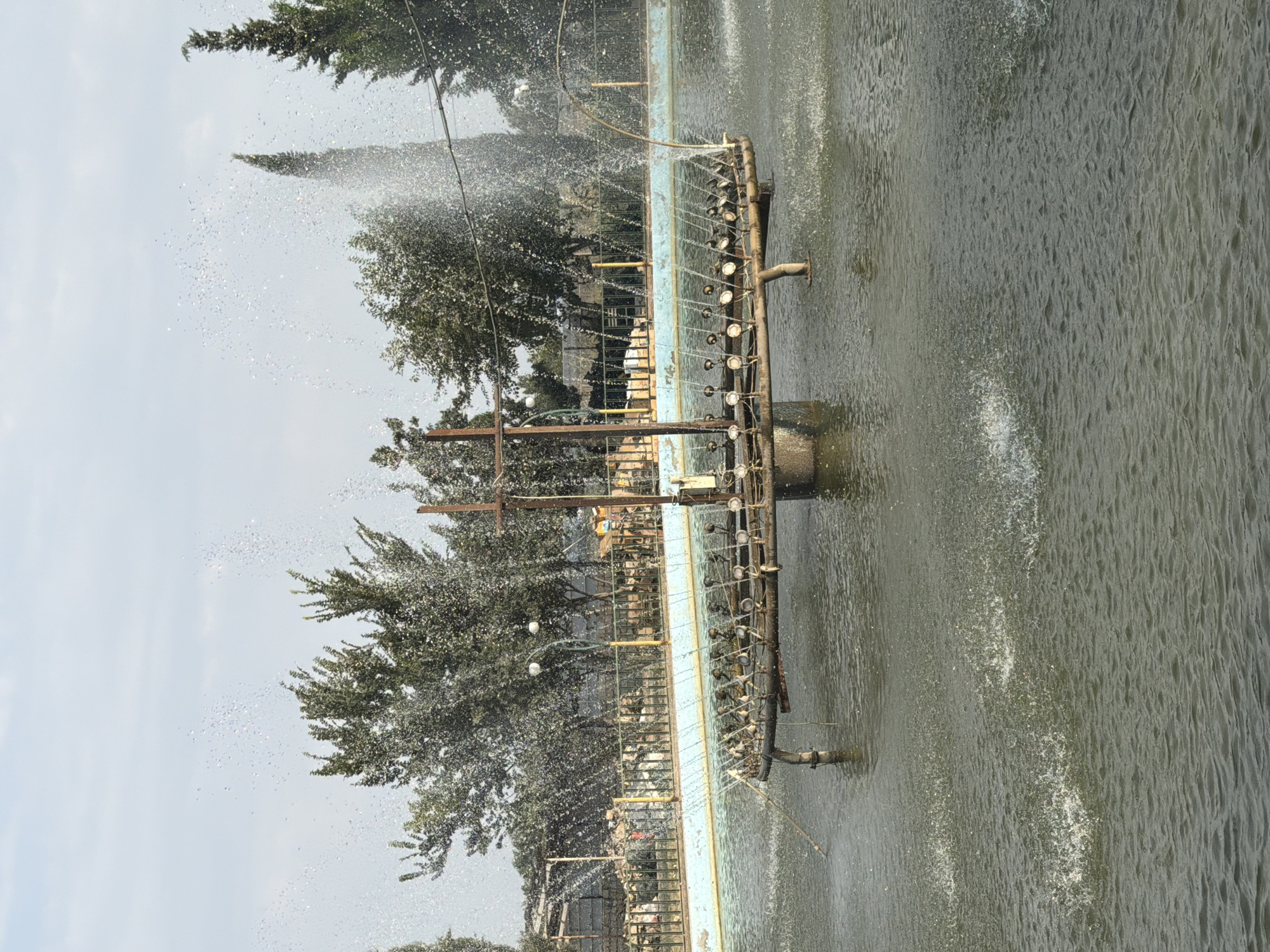 The image shows a close-up view of a fountain in a body of water, likely in the city of Nəvahi Stansiya, Azerbaijan. The fountain has a complex structure, resembling a ship's deck with multiple nozzles spraying water upwards. The water in the fountain is choppy and agitated due to the spraying. In the background, there are trees and a fence, suggesting an outdoor setting, possibly a park or a public area. The sky is visible, with some clouds. There are no people visible in the image.