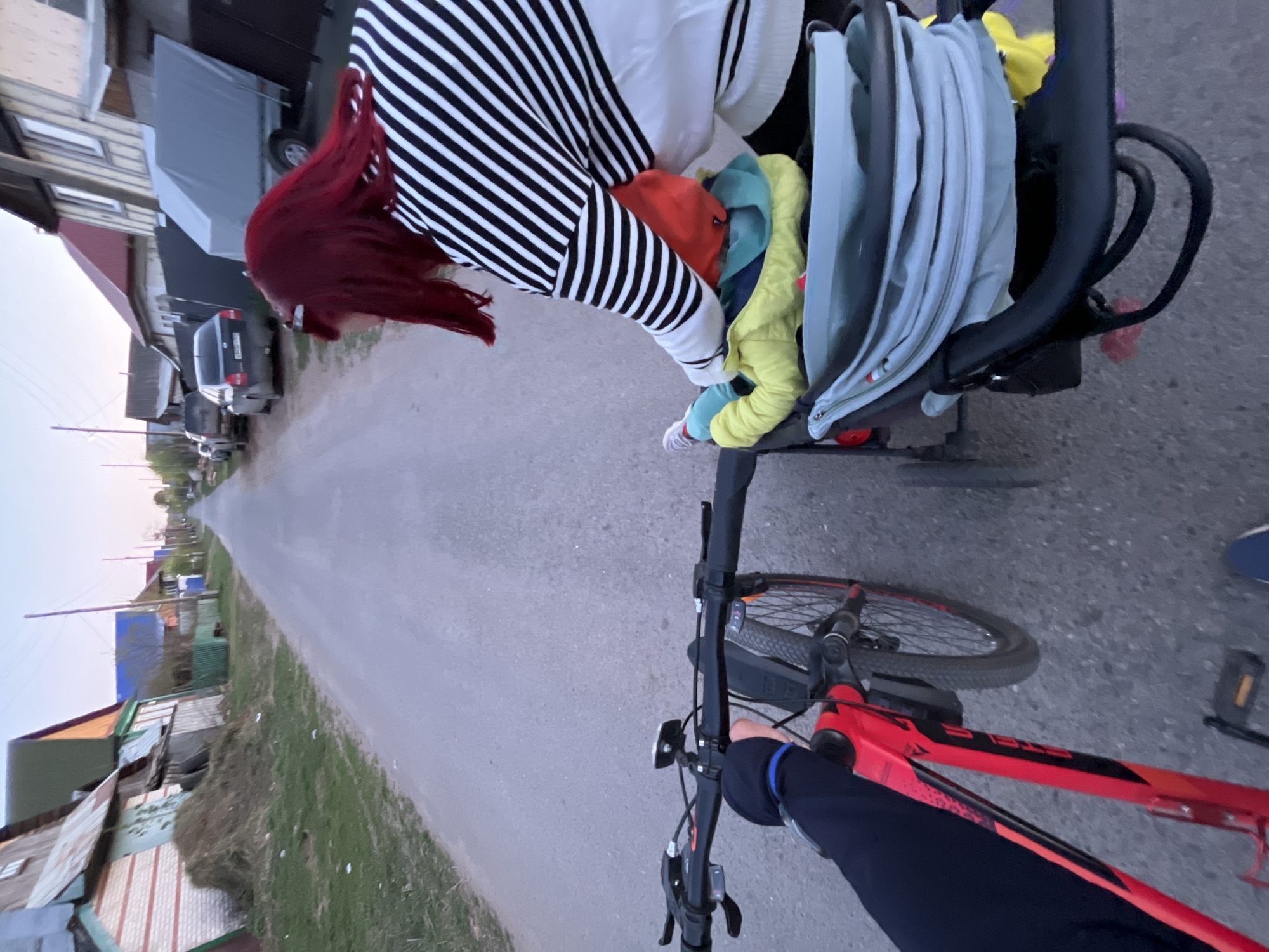A first-person perspective photograph depicts a bicycle being ridden down a narrow asphalt road in what appears to be a residential area in Chaikovsky, Russia. The rider is wearing a dark blue long-sleeved shirt.
To the right and slightly in front of the bicycle is a baby stroller with what appear to be blankets and clothing inside. A woman with bright red hair, wearing a white sweater with black horizontal stripes, is bent over the stroller, possibly interacting with a child inside.
The road extends into the distance, lined with modest, single-story homes on either side, many with corrugated metal roofing. Several vehicles are parked along the roadside further up the street. The sky overhead is pale and overcast.
