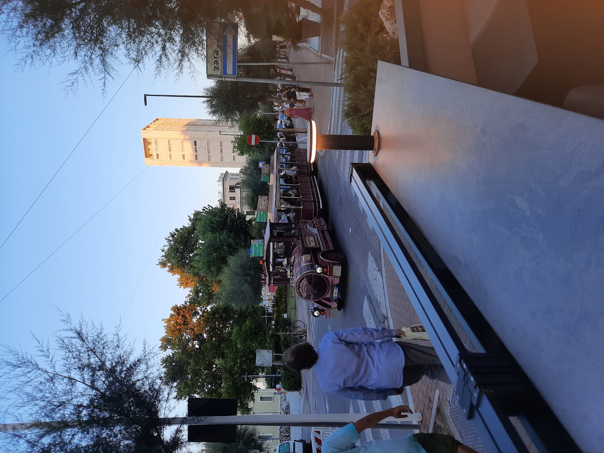 In Pesaro, Italy, at dusk, a tourist train resembling a vintage locomotive travels along a street lined with trees and buildings. The train, adorned with ornate details and illuminated, carries passengers. In the foreground, a person in a blue shirt walks away from the camera, carrying a shopping bag. On the right side of the street, a Zara sign is visible above a pathway where pedestrians stroll. A tall, modern building stands in the background, catching the warm light of the setting sun. The scene suggests a leisurely evening in a coastal Italian city.