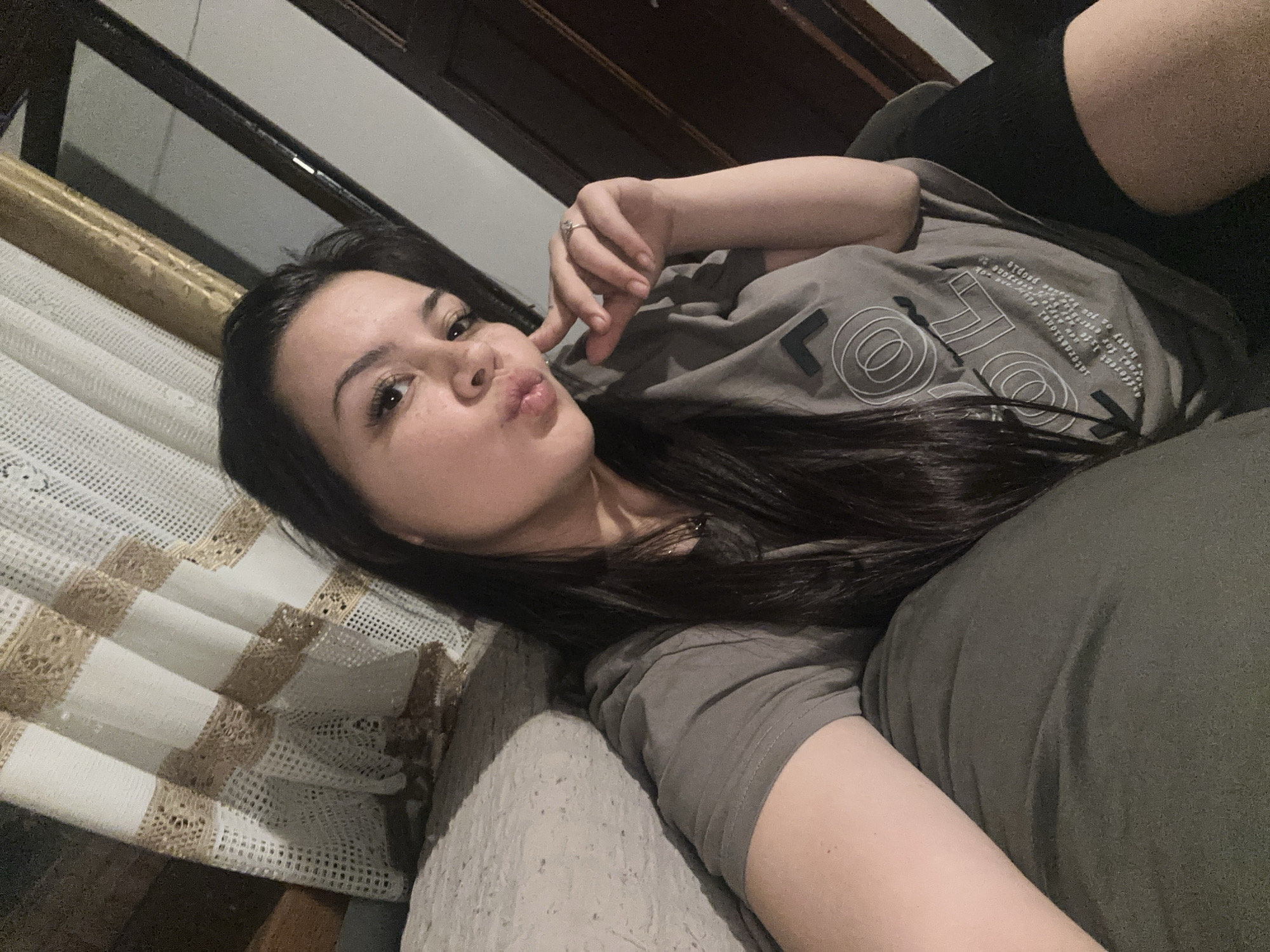 A young woman is shown in a close-up, chest-up selfie, lying down in an indoor setting, potentially a home in Chubra, Bulgaria. She has long, dark hair and is looking directly at the camera with a playful, pouty expression. Her left hand is raised, with fingers near her face, and a ring is visible on her left ring finger.
She is wearing a grey t-shirt with white graphic designs and text, which includes partially visible words such as "people," "different," "happy," and "yourself," along with circular and angular motifs. She also appears to be wearing black leggings or high socks. The woman is resting on a surface that resembles a sofa or bed, covered with a textured, light-colored fabric and an olive-green cushion or throw. To her left, a white and beige crocheted or lace textile is draped over a piece of furniture. The background features dark wooden elements and a light-colored wall, indicating a domestic interior. The lighting suggests an indoor setting, possibly during the evening or night.